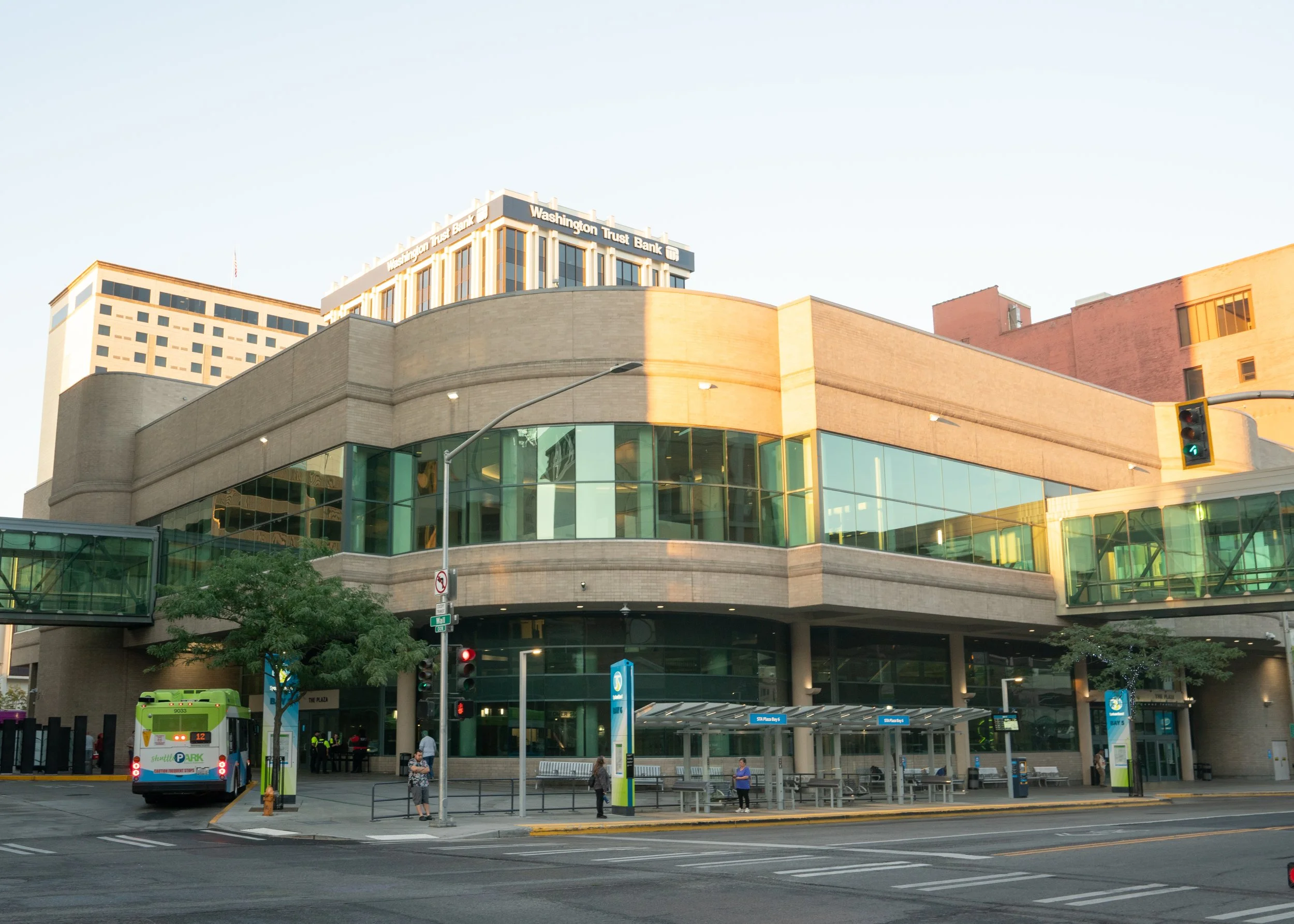 Photo of a city street scene showing a modern building with a rounded corner and large glass windows, a green shuttle bus, traffic lights, pedestrians, and nearby buildings. The building has a sign that reads 'Washington Trust Bank'.