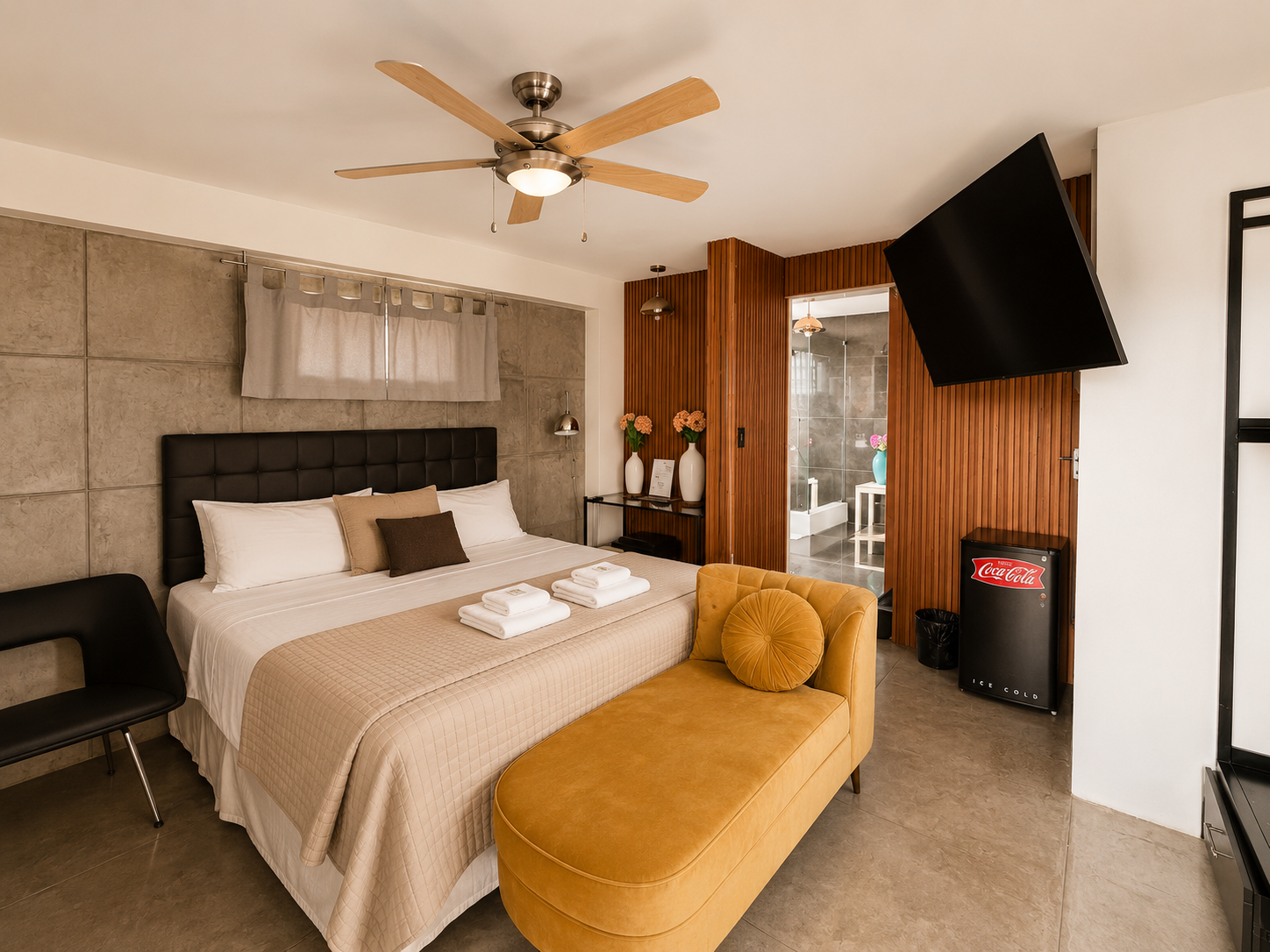 Hotel room with a bed, black headboard, beige bedspread, yellow bench at foot of bed, TV mounted on wall, small fridge, and bathroom with shower in background.