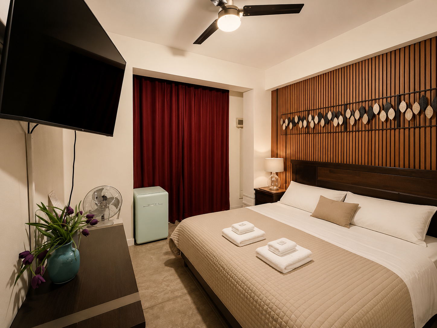 Hotel room with a king-sized bed, beige quilt, and pillows, wooden headboard, wall decoration of hanging leaves, side table with lamp, red curtains, hanging on the wall TV, small fan, air purifier, and vase with purple tulips.