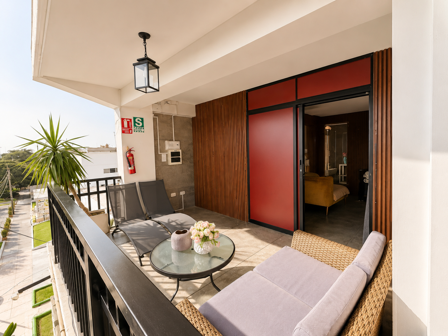 Balcony with black railing, lounge chairs, a wicker sofa with cushions, a glass-top coffee table with a flower vase, potted plant, and a view of neighboring buildings and greenery.