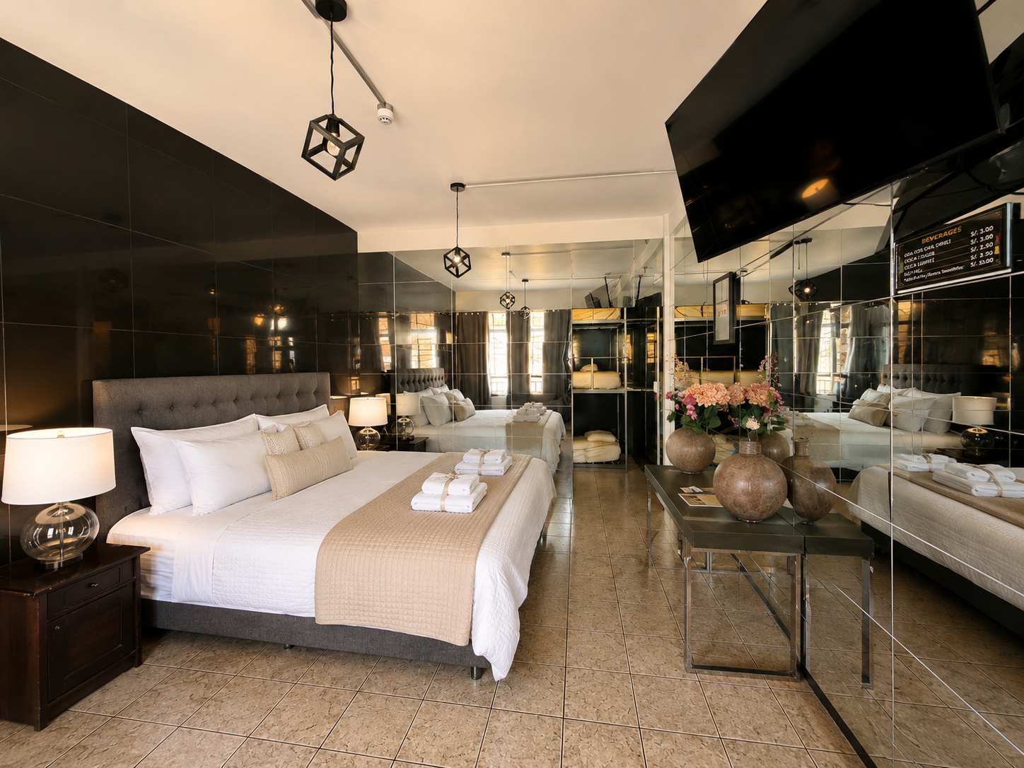 A modern hotel room with a large bed, bedside lamps, a mirrored wall, and decorative vases with flowers.