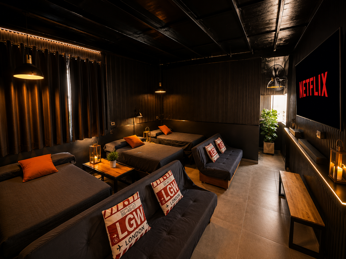 A cozy, modern bedroom with four beds, black bedding, and orange pillows. The room has dark walls and curtains, with warm lighting from hanging lamps and lanterns. There is a flat-screen Netflix TV on the wall and a bench at the foot of the beds, along with some indoor plants and decorative candles.
