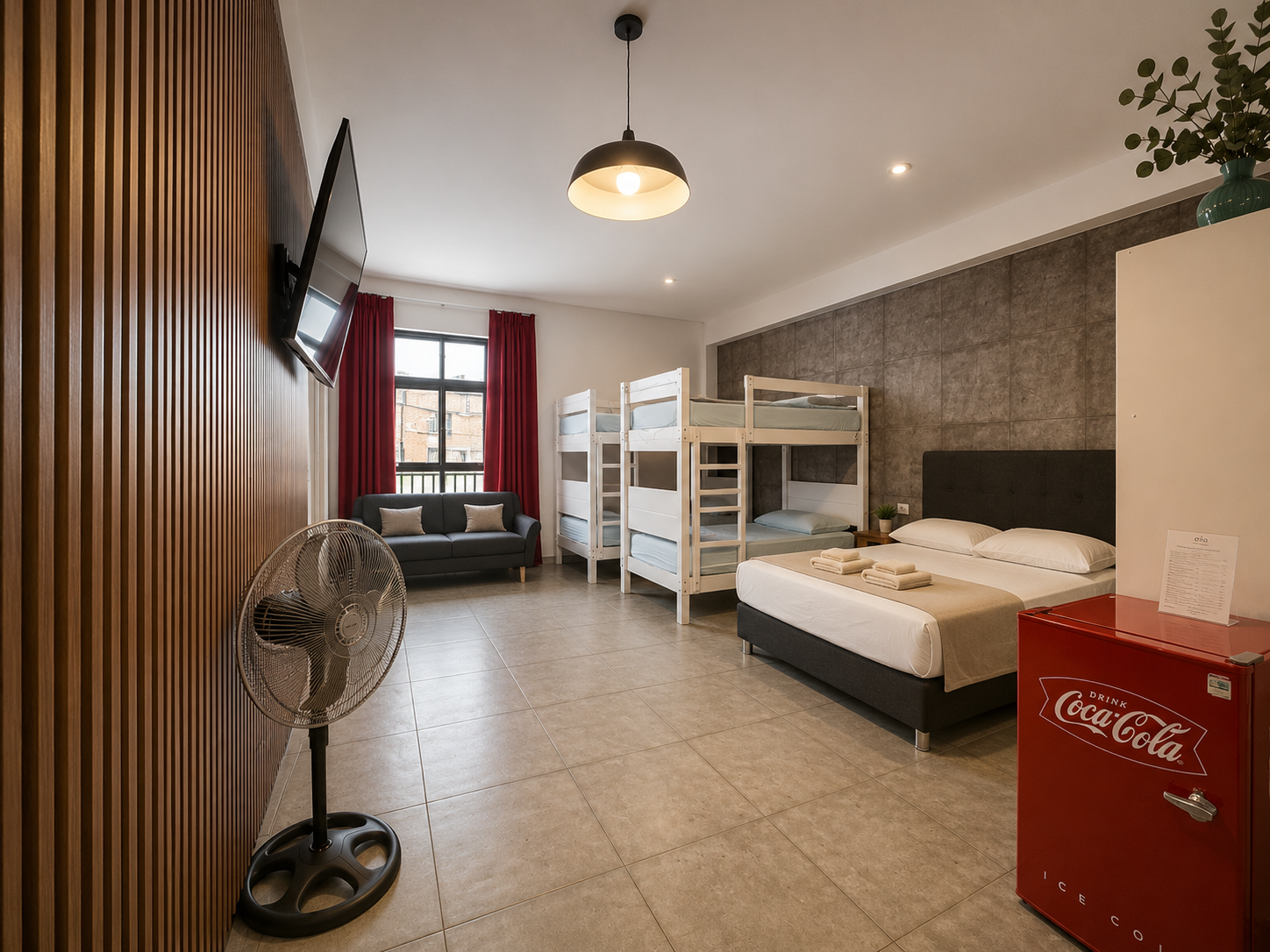 Hotel room with a double bed, bunk beds, a black sofa, a fan, a TV on the wall, red curtains, a window with city view, and a red Coca-Cola fridge.