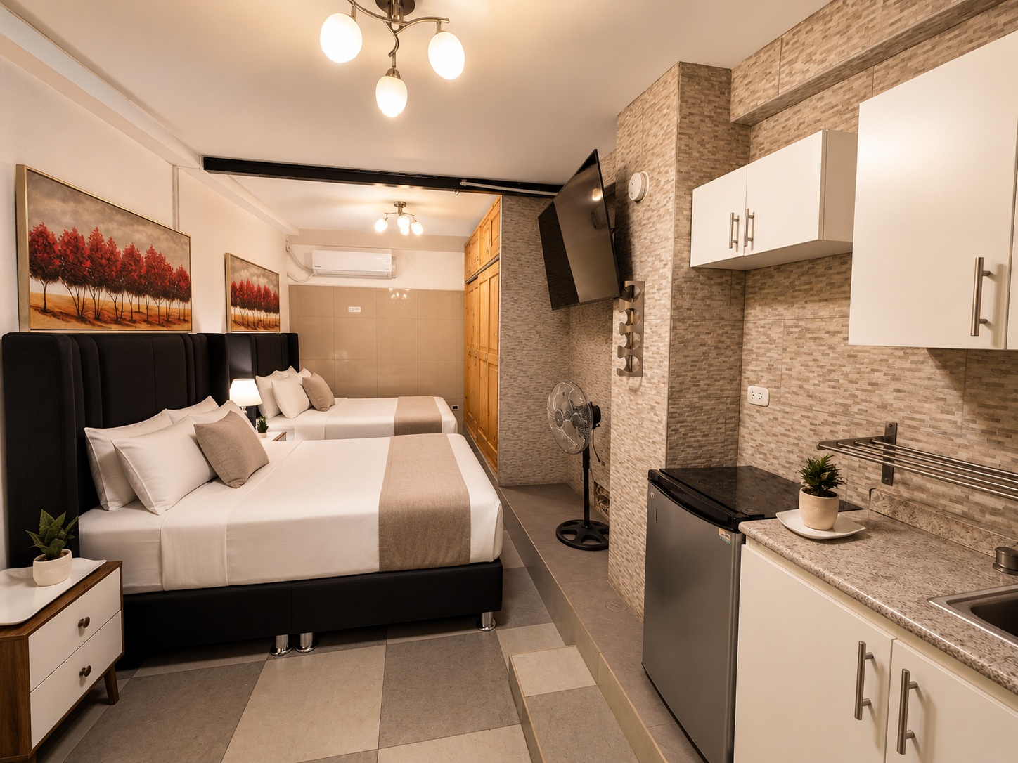 Hotel room with two beds, headboards, artwork, small nightstand, lamp, and a kitchenette with a mini fridge, sink, cabinets, and decorative plants, with a wall-mounted TV, fan, and air conditioning