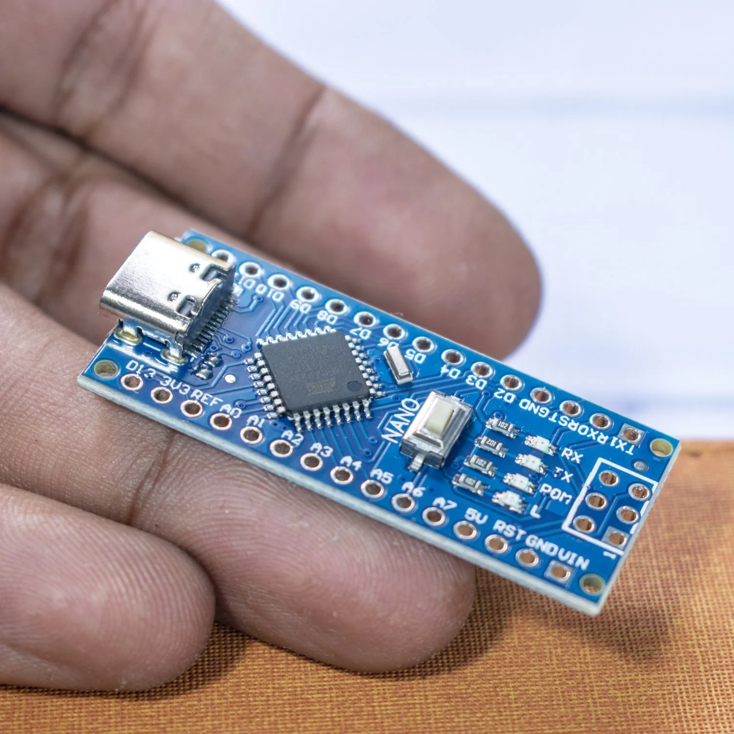 Close-up of a blue Arduino Nano microcontroller board held between fingers, showing various electronic components and pins.