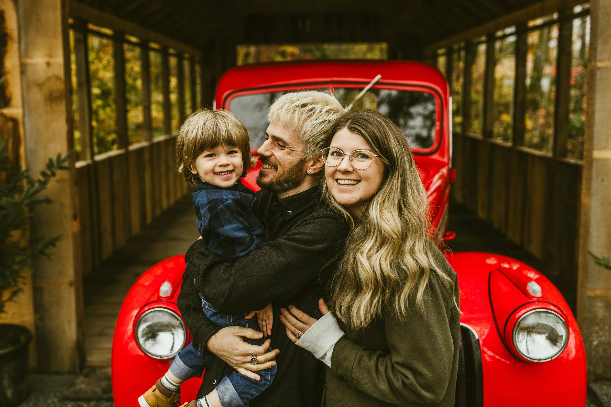 Family of three hugging beside a vintage red truck on a wooden bridge