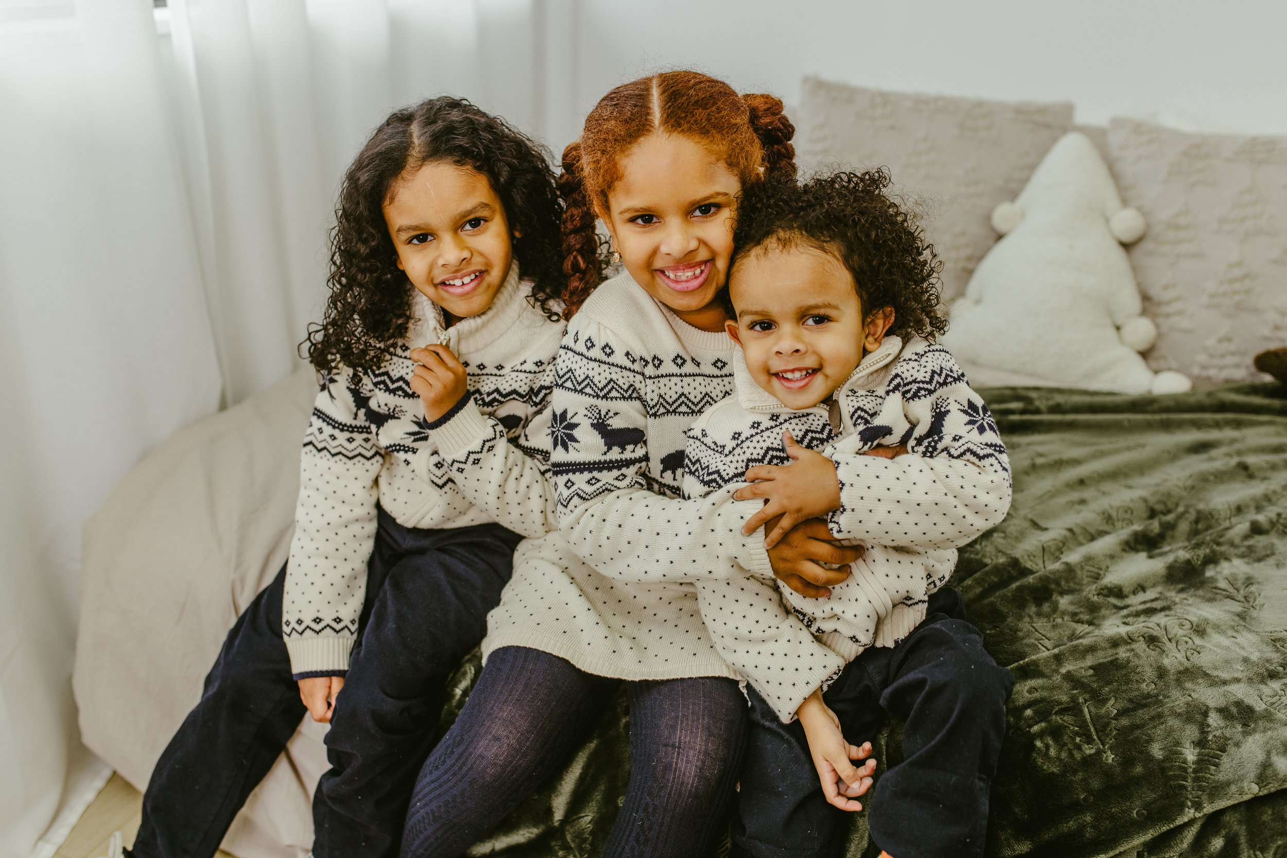 Three sisters in matching reindeer sweaters, close together on a bed