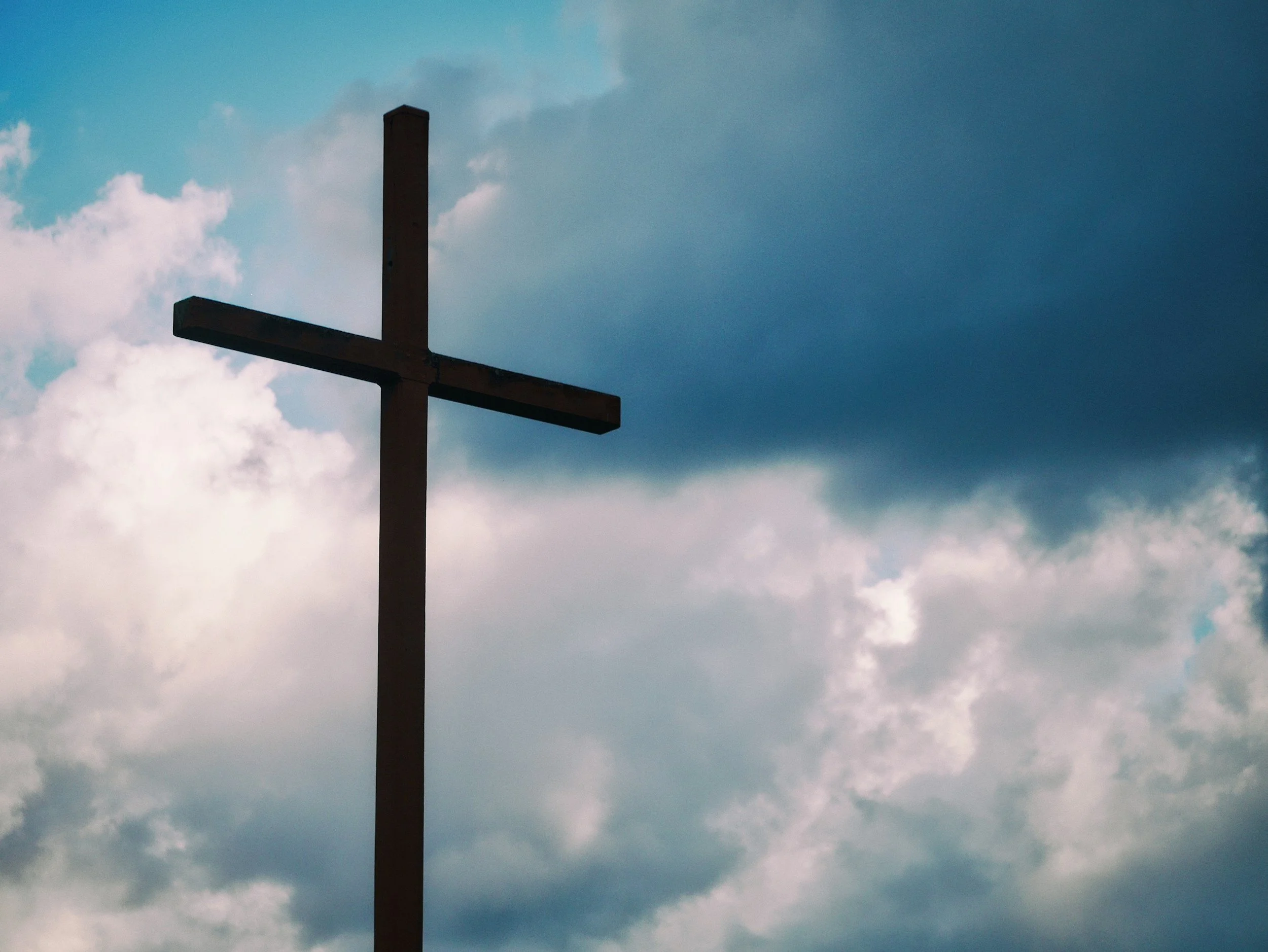 A silhouette of a cross against a sky with clouds and a gradient of blue hues.