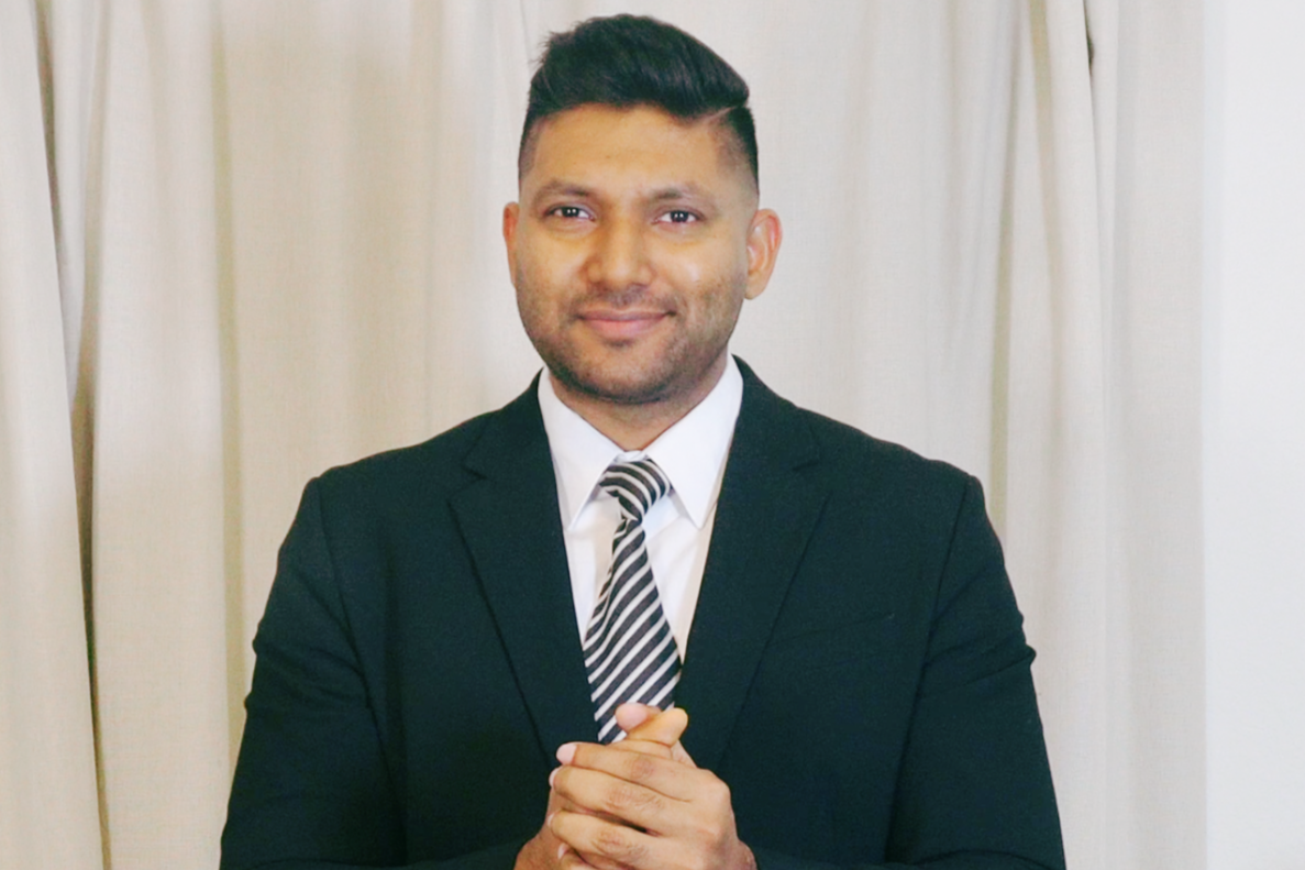 A man in a black suit with a white shirt and striped tie, standing in front of cream-colored curtains, smiling with hands clasped.