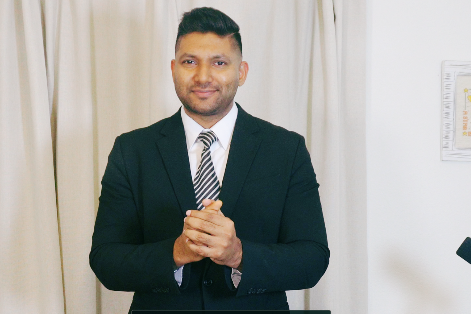 A man in a business suit with a striped tie standing in front of cream-colored curtains and a white wall, smiling and clasping his hands together.