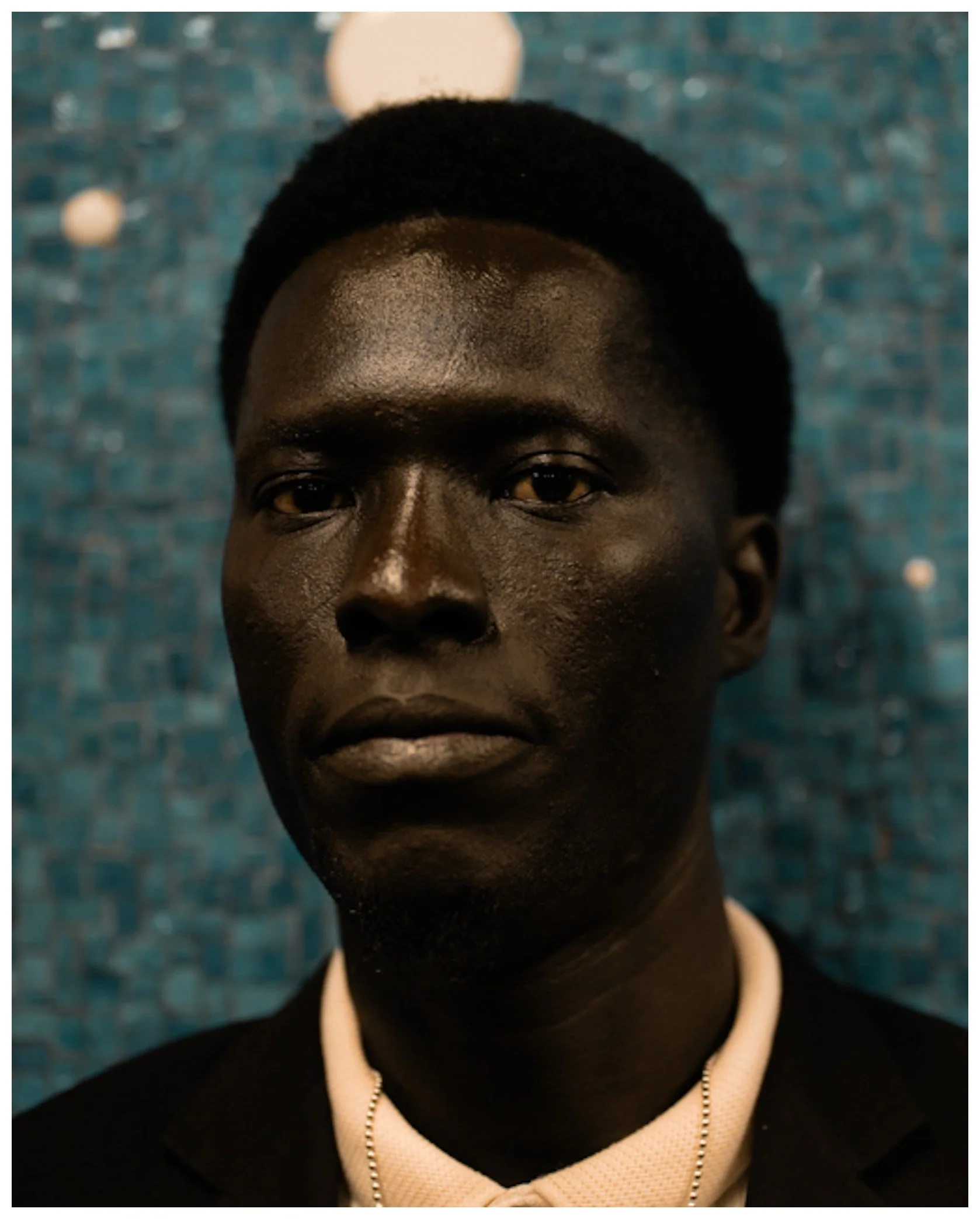 Close-up of a person with dark skin and short black hair, wearing a black shirt, beige hoodie, and a pearl necklace, against a blue mosaic background with white dots.