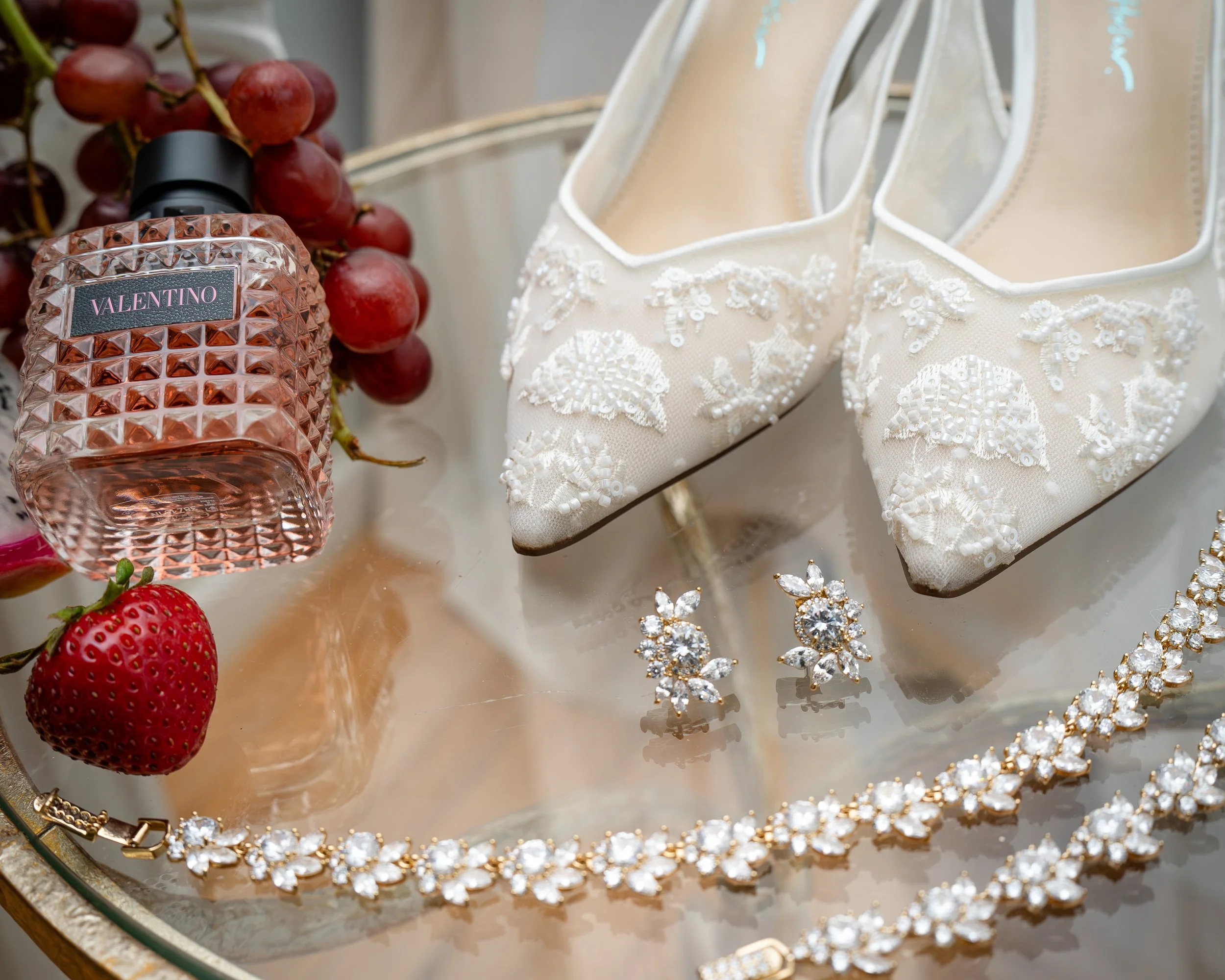 A collection of red grapes, a bottle of Valentino perfume, a pair of white embroidered shoes with pearl and sequin details, strawberries, and jewelry including earrings and a necklace with large white stones.