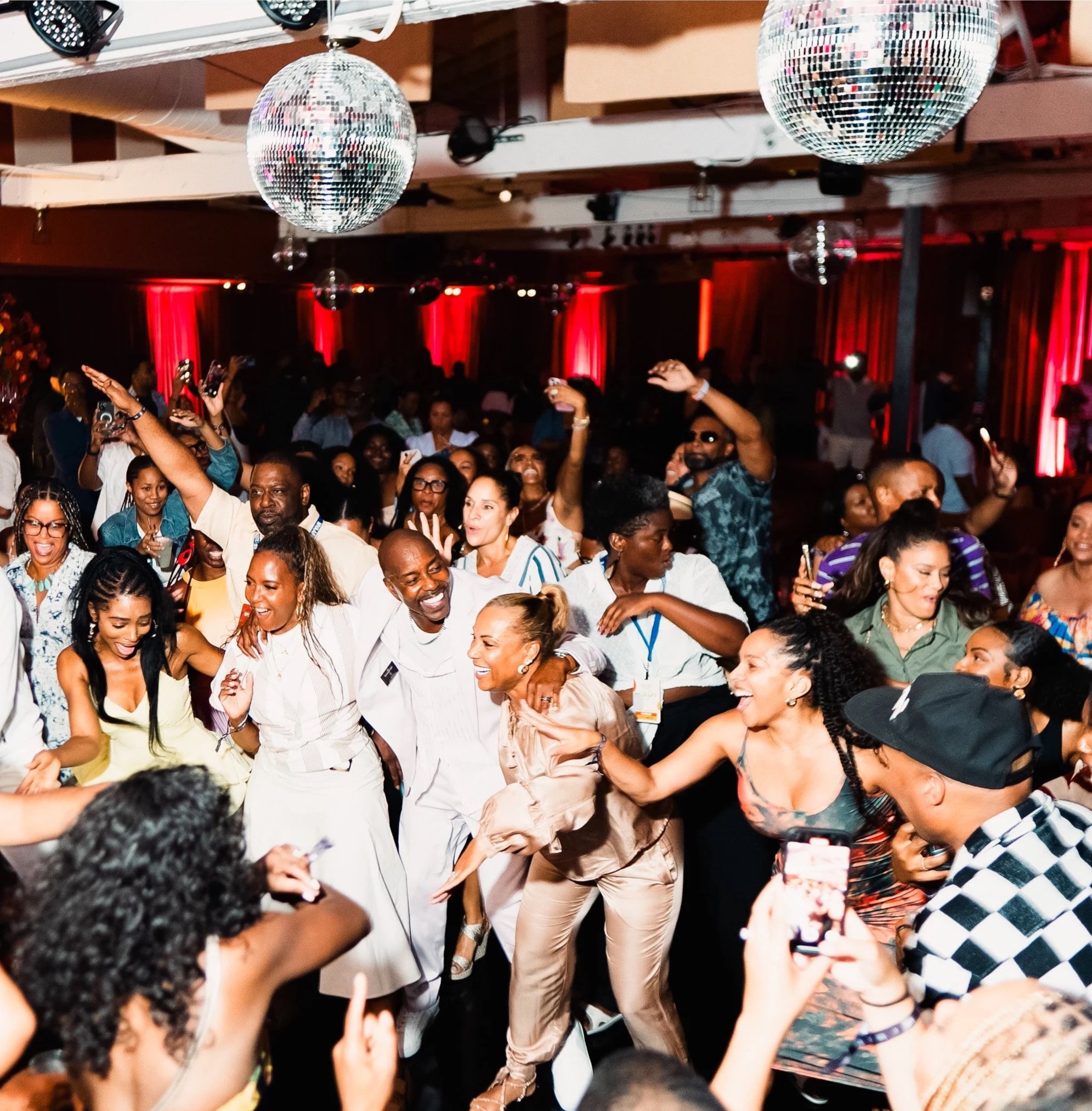 A lively event with many people dancing and enjoying themselves under disco balls with red lighting in the background.