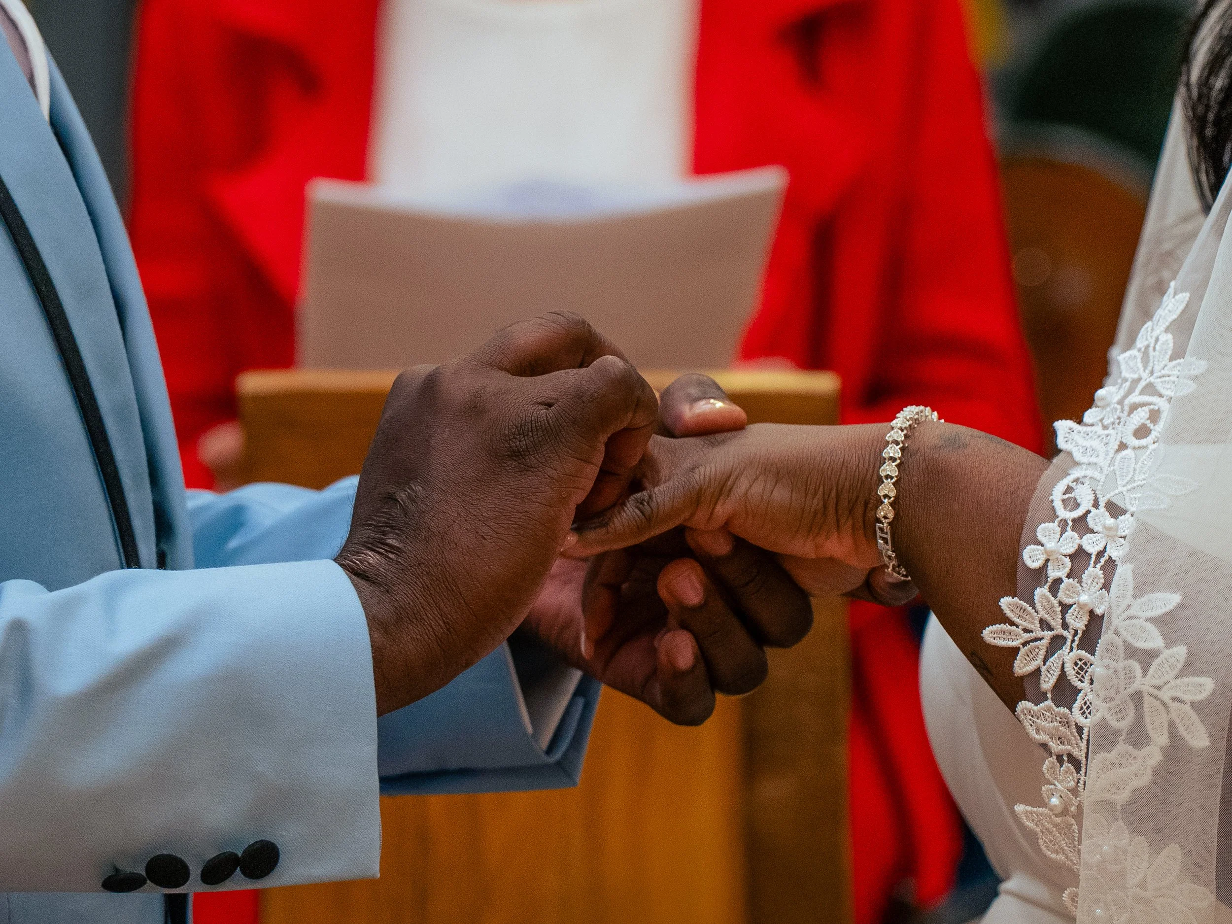A close-up of a couple holding hands during a wedding ceremony. The man is dressed in a light blue suit, and the woman is wearing a white lace dress and a delicate bracelet. An officiant in a red jacket is visible in the background.