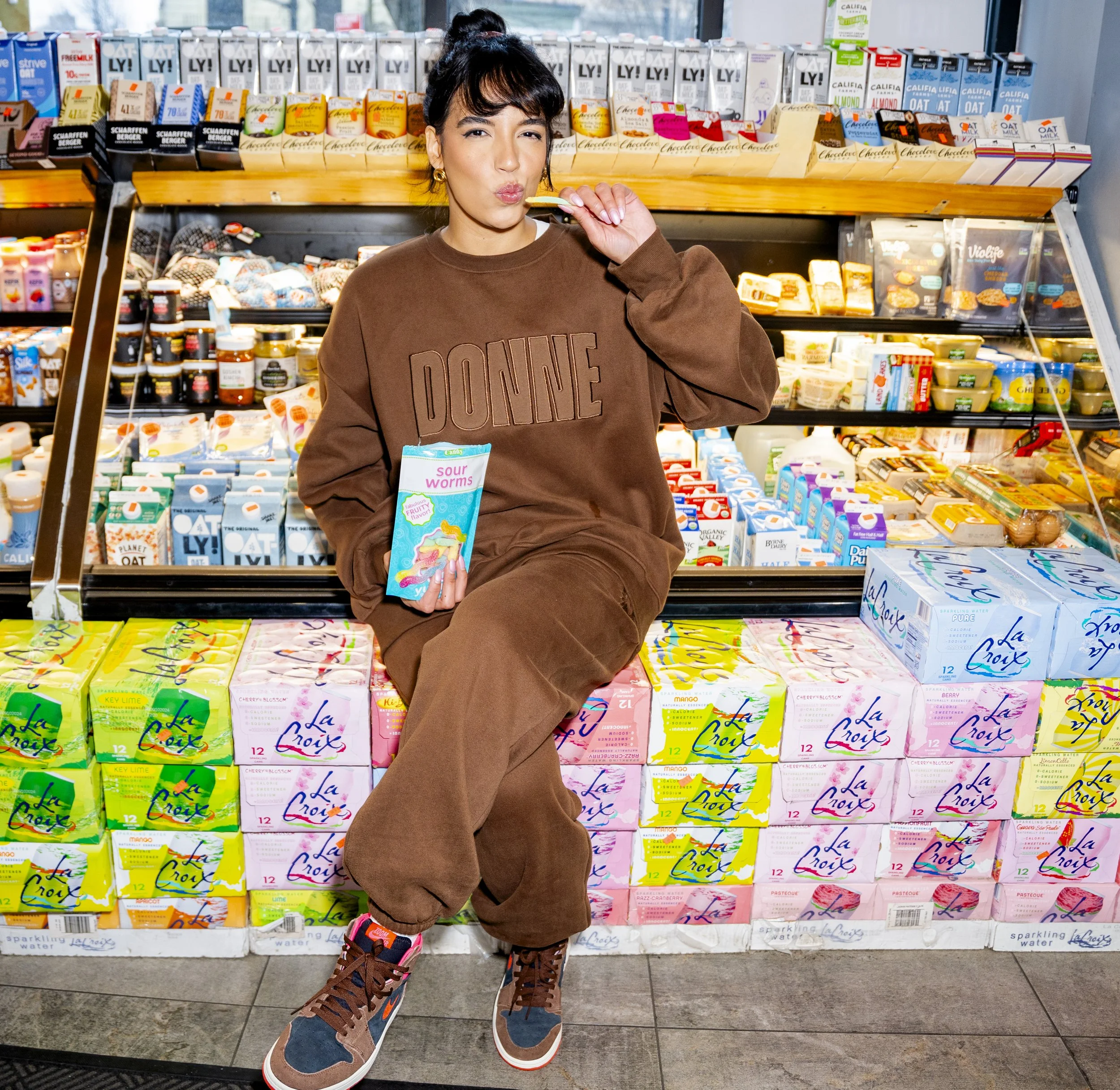 A young woman with dark hair, wearing a brown sweatshirt with 'DONEY' printed on it, sitting on a store display in front of a shelf stocked with various cartons of flavored and flavored water. She is holding a sour worms candy in one hand and winking at the camera.