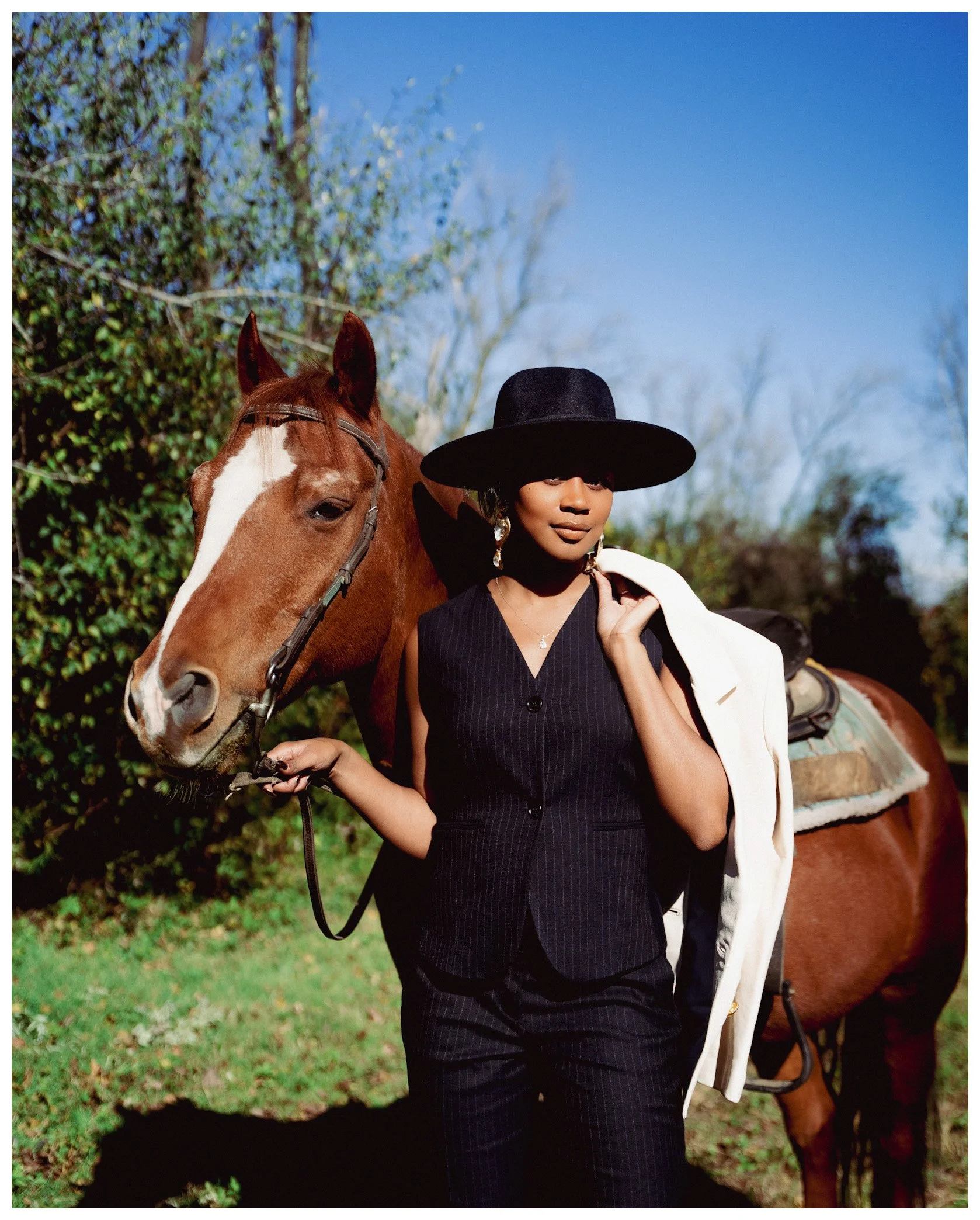 A woman with dark hair wearing a black wide-brimmed hat, black sleeveless dress, and carrying a white jacket over her shoulder, holding the reins of a brown horse with a white blaze on its face, in an outdoor setting with trees and greenery.