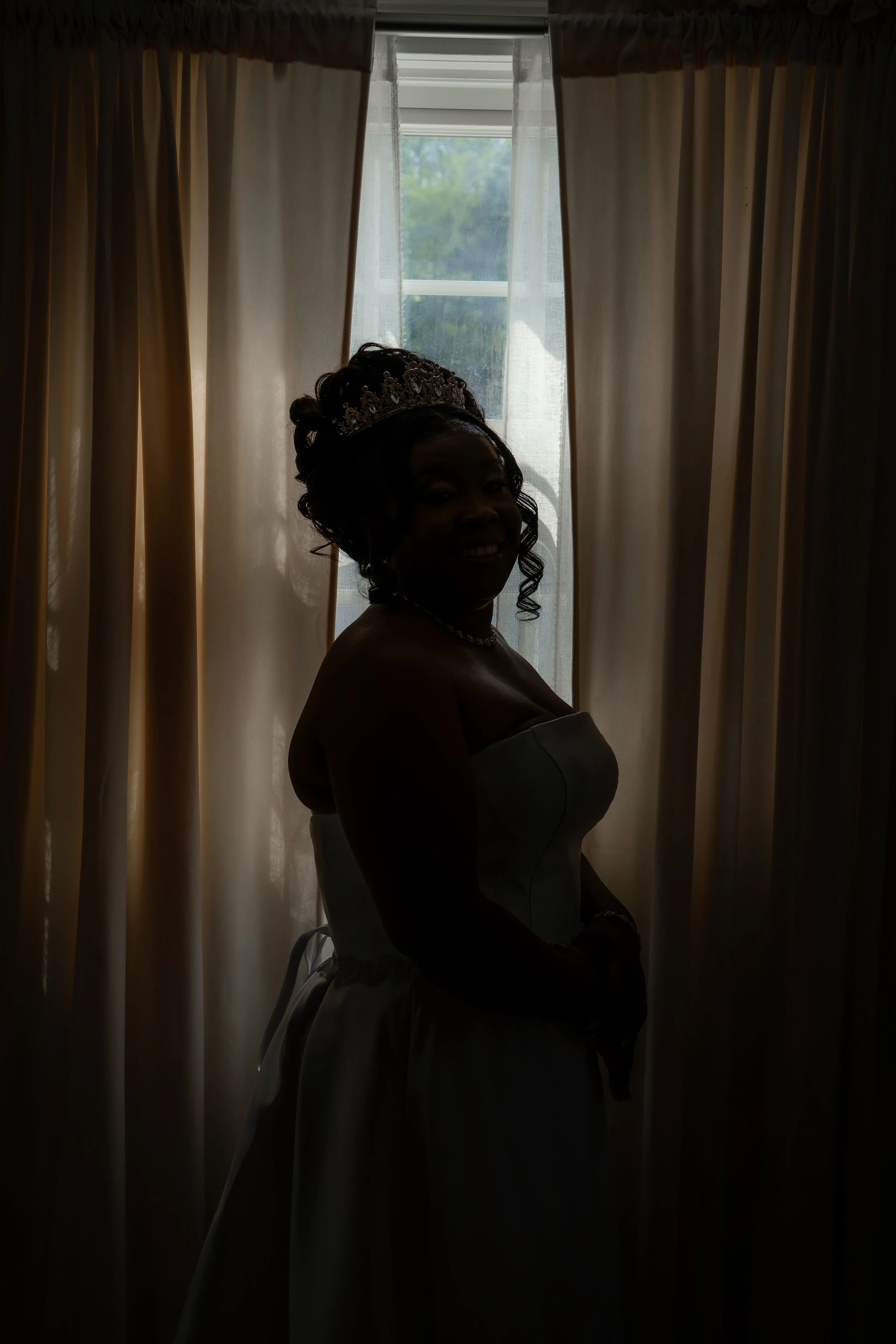 Silhouette of a woman in a wedding dress with an ornate tiara, standing in front of a window with light-colored curtains, with a partly visible smile.