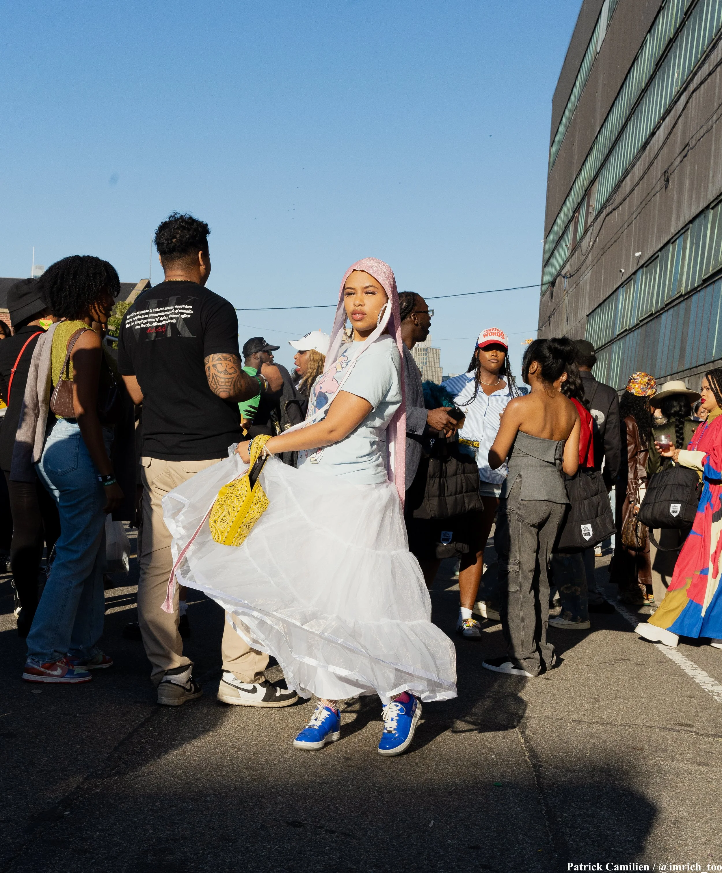 A young woman wearing a pink headscarf and a white dress with a ruffled bottom, standing among a crowd of diverse people outdoors on a sunny day, with tall buildings in the background.