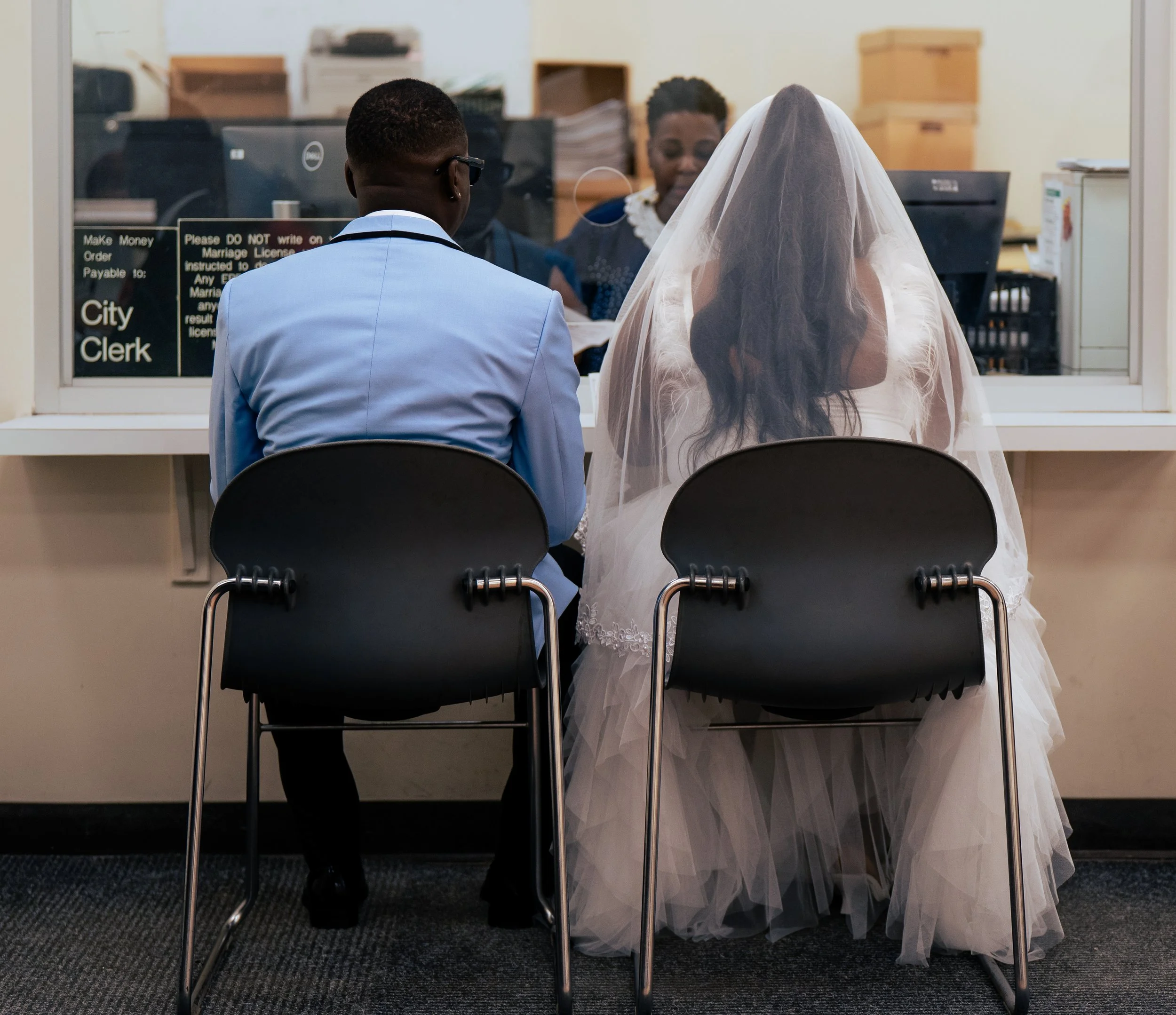 A couple at a wedding sitting at a counter in a government office, with a woman behind the glass window processing paperwork.