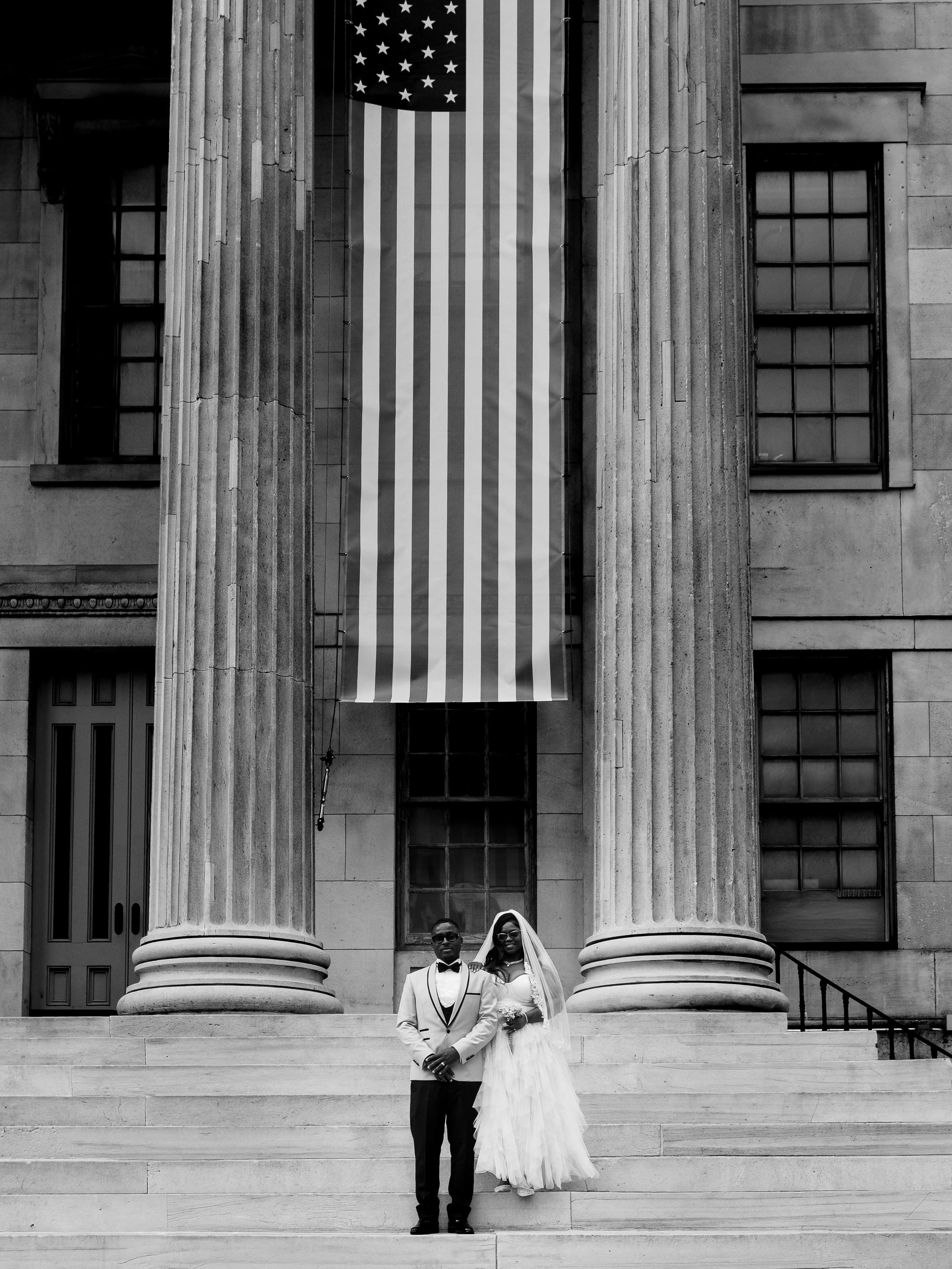 A newlywed couple standing on steps in front of a government building with large columns and an American flag hanging between them.