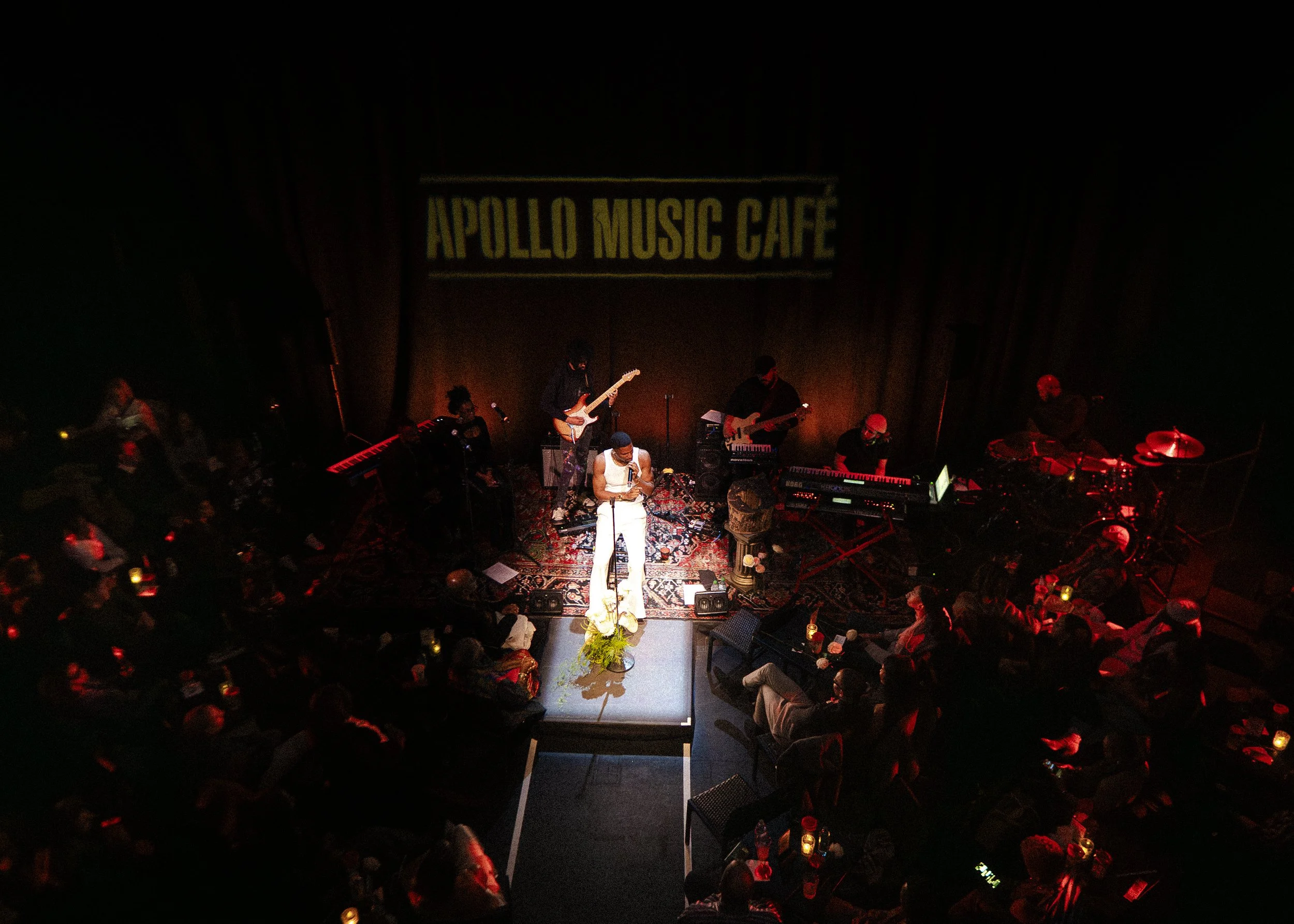 A live music performance at Apollo Music Cafe with musicians playing guitar, keyboard, and drums on stage, surrounded by an audience with dim lighting and candles.