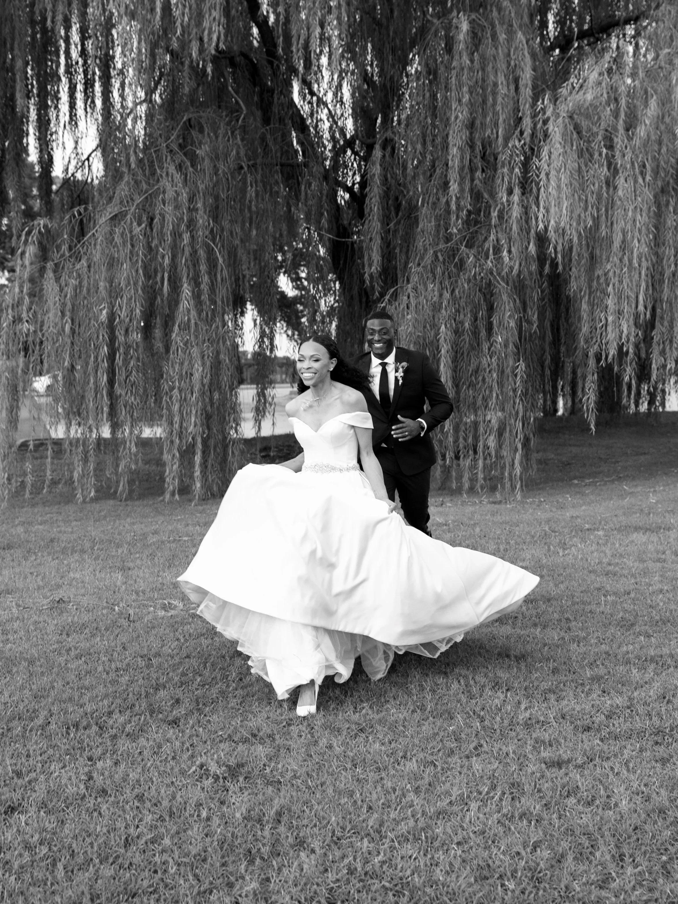 A bride and groom are dancing outdoors on a grassy area with trees in the background. The bride is wearing a white wedding dress and the groom is in a black suit with a boutonniere, both smiling joyfully.
