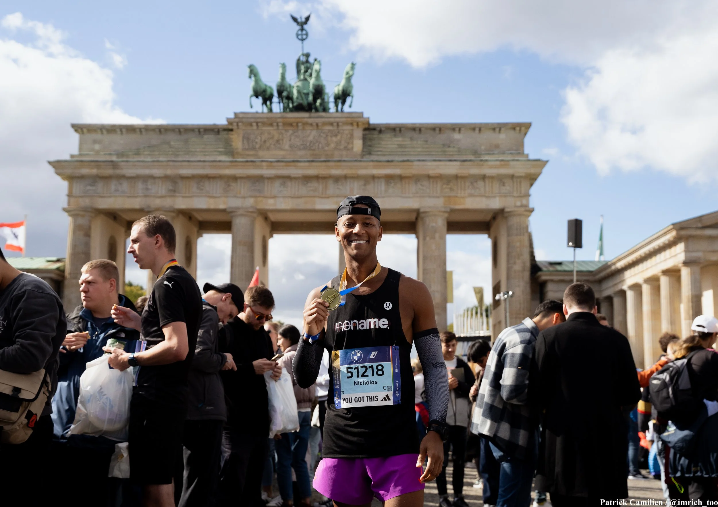 A smiling male marathon runner named Nicholas holding a finisher medal at the Berlin Marathon with the Brandenburg Gate in the background on a partly cloudy day.