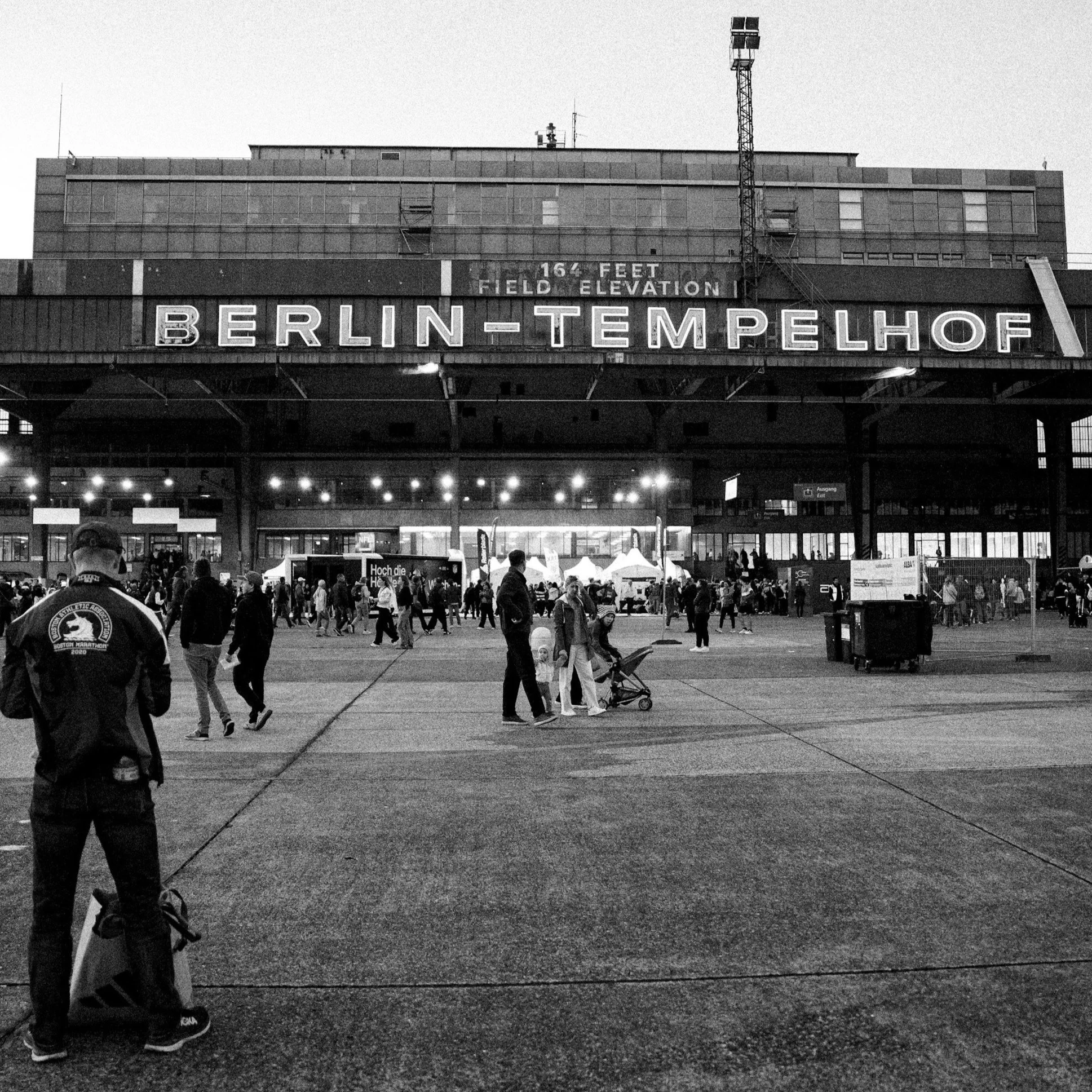 Black and white photo of the Berlin-Tegel airport terminal. A large sign displays 'BERLIN-TEMPELHOF'. The building has a tall tower with speakers or lights on top. People are walking outside the terminal, some with luggage, and some are gathered near small tents or booths.
