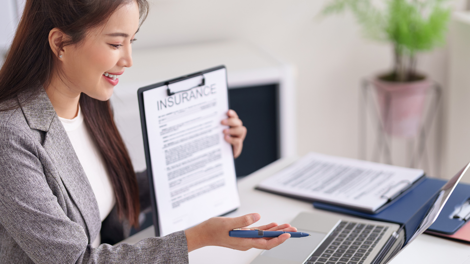 Smiling woman in business attire holding a clipboard with an insurance document, sitting at a desk with a laptop, pen, and paperwork in an office.