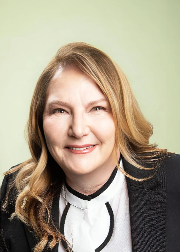 A woman with shoulder-length wavy blonde hair smiling at the camera, wearing a white blouse with black accents and a black blazer, in front of a light green background.