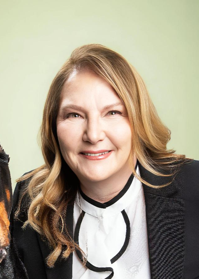 A woman with wavy light brown hair wearing a black blazer and a white blouse with black trim, smiling against a light green background.