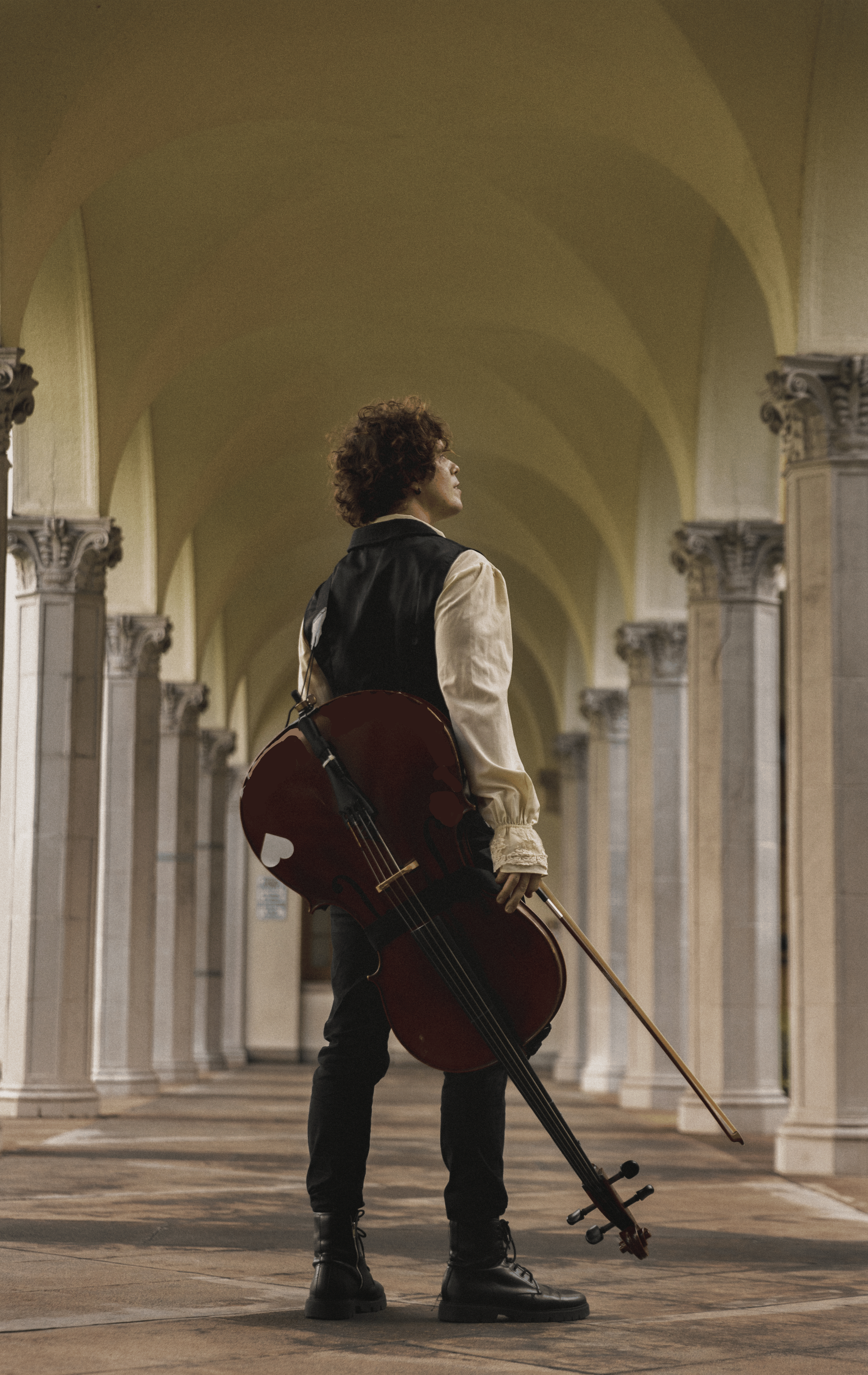Lovejack dressed in a beige shirt and black vest, holding a cello and bow, standing in a grand hallway with tall, ornate columns and vaulted ceilings.