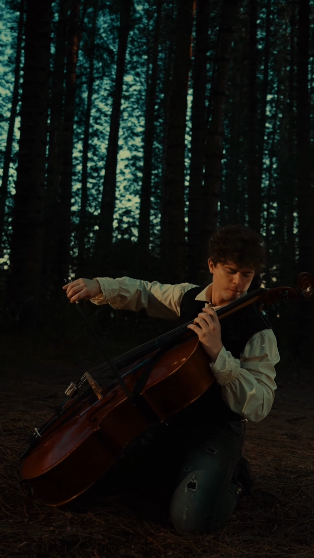 Lovejack playing cello in a forest while wearing a black vest and ruffled white shirt.