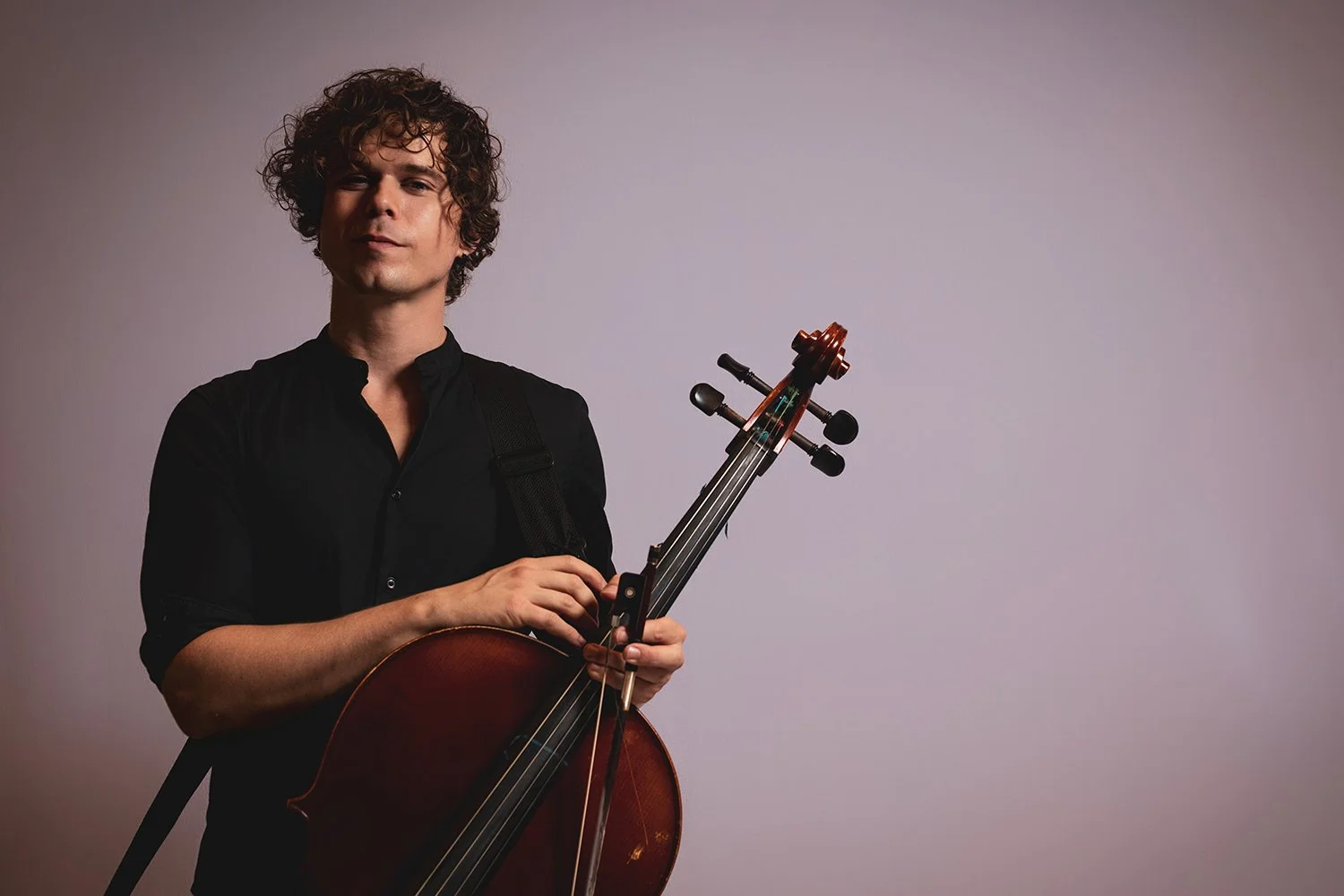 A young man with curly hair wearing a black shirt holding a cello and bow against a plain background.