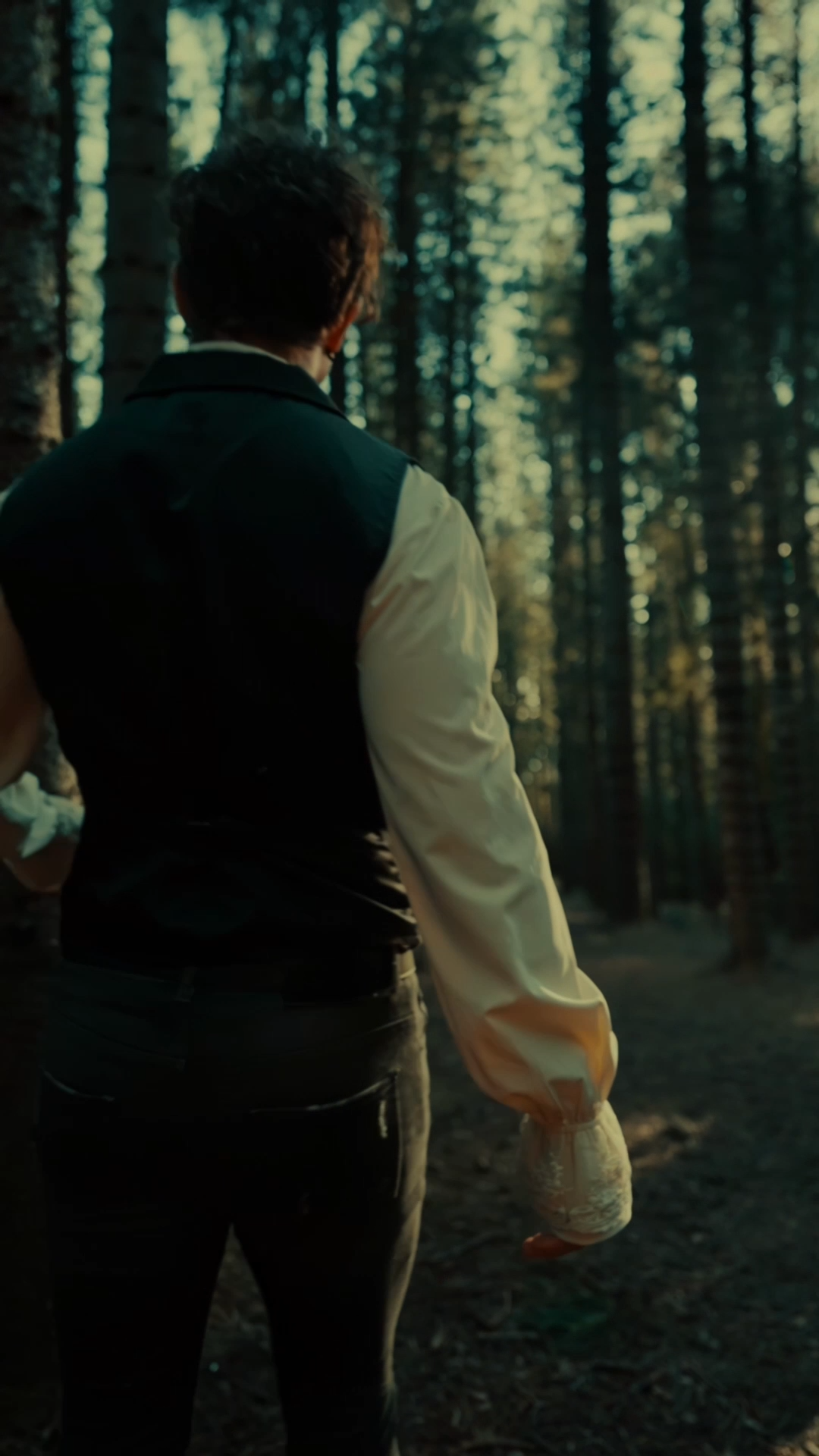Lovejack wearing a black vest and cream-colored sleeves, walking through a dense forest with tall trees and sunlight filtering through.