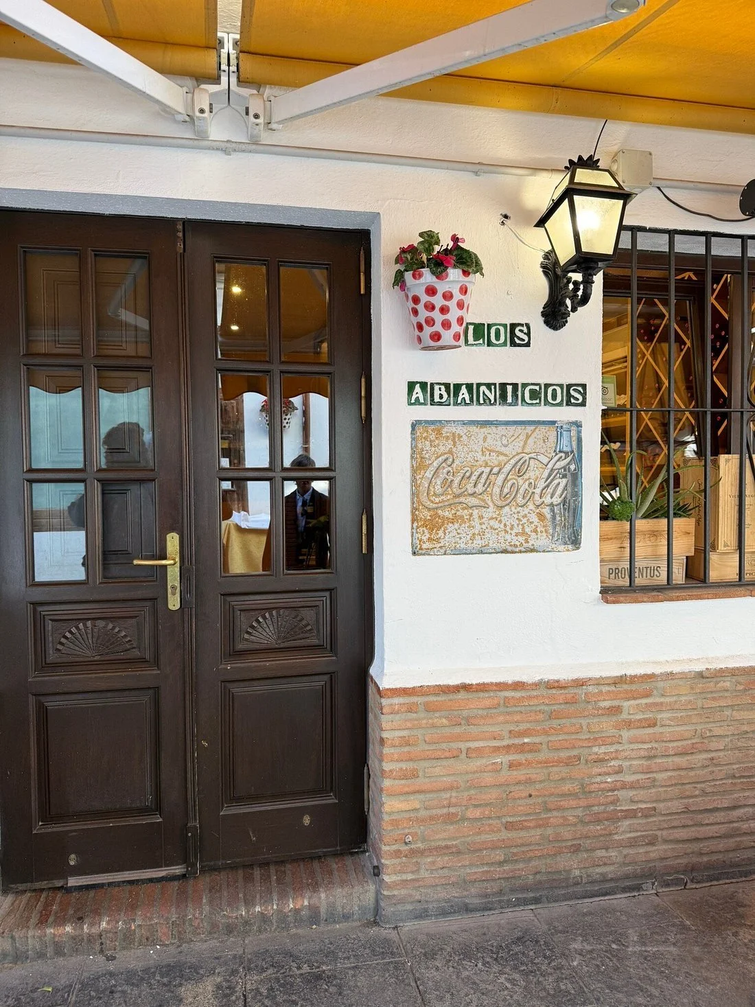 Entrada de un restaurante con cartel que dice "Los Abanicos" y un cartel de Coca-Cola. Ventana con rejas y plantas decorativas.