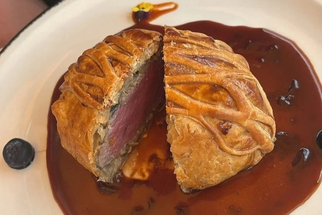 Pastel de carne en forma de media luna con corte que muestra carne cocida en medio, rodeado de salsa oscura con algunas bayas negras y decoraciones de flores.