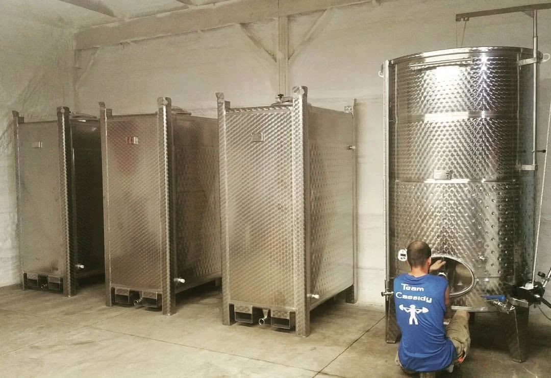 A person in a blue shirt kneeling and working on the large hatch of a tall stainless steel fermentation tank in a brewery, with four smaller stainless steel tanks lined up against the wall in the background.