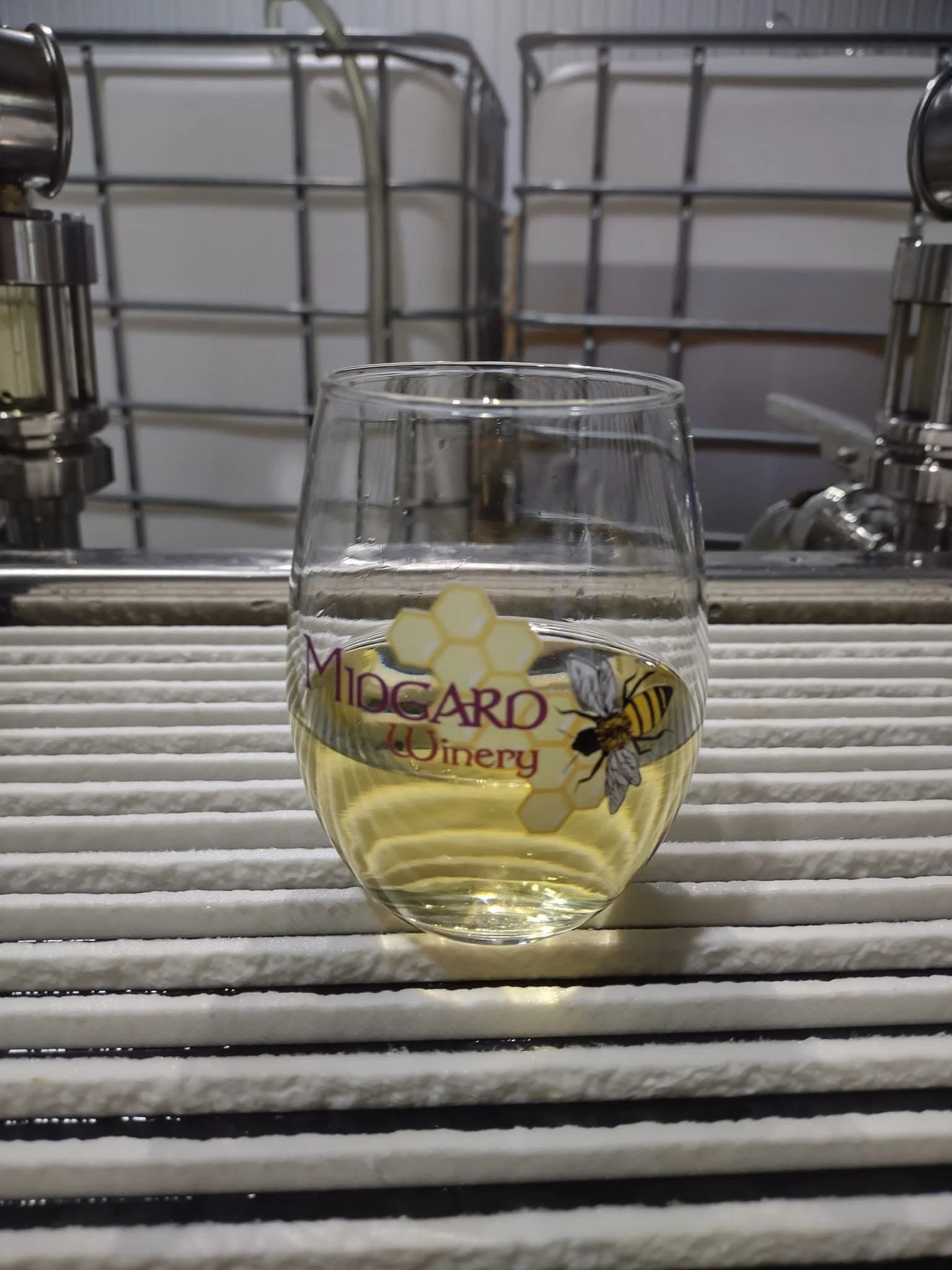 A glass of white wine with a label reading 'Midgard Winery' and a bee illustration, placed on a white slatted surface in front of industrial kitchen racks.