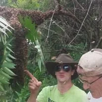 Two people in a lush, green outdoor setting with tropical plants. One person is wearing a large hat and sunglasses, and the other is wearing a cap, both partially visible.