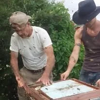Two men outdoors, one wearing a baseball cap and glasses, the other wearing a wide-brimmed hat, examining or working on a glass-covered frame or surface.