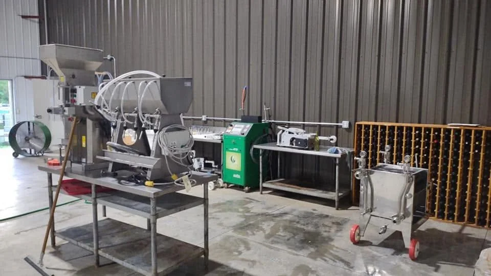 Industrial equipment with a metallic framework, piping, and control panels set up on a concrete floor inside a warehouse or factory. There is a large metallic machine on the left, a green control unit in the middle, and a stainless steel chamber on wheels to the right, with a yellow rack containing small bottles or containers behind it.