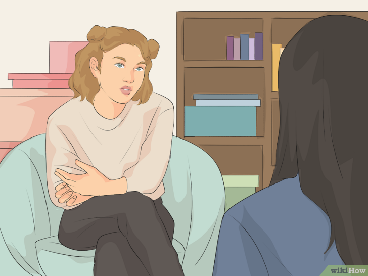 A young woman with light brown hair styled in buns, sitting on a light blue chair, talking to another person with long black hair, in a room with a bookshelf and pink boxes in the background.