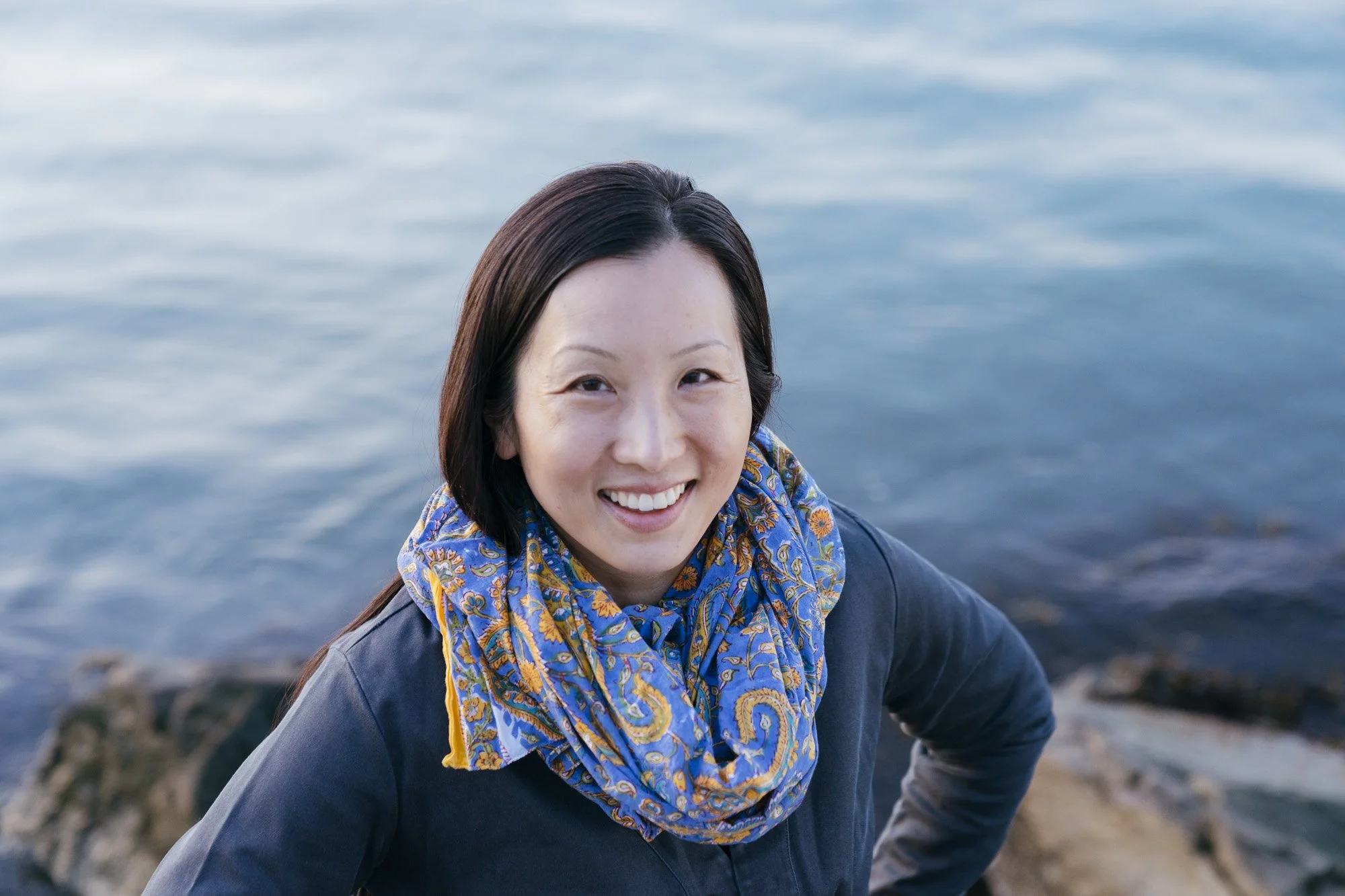 A smiling Asian American woman stands outdoors near water, wearing a blue patterned scarf and dark jacket.