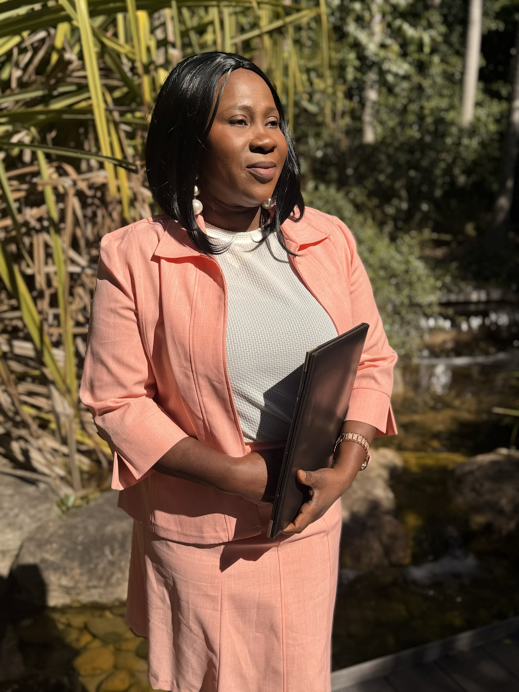 A woman with black hair wearing a peach blazer and matching skirt, holding a folder, standing outdoors near a pond with plants and rocks.