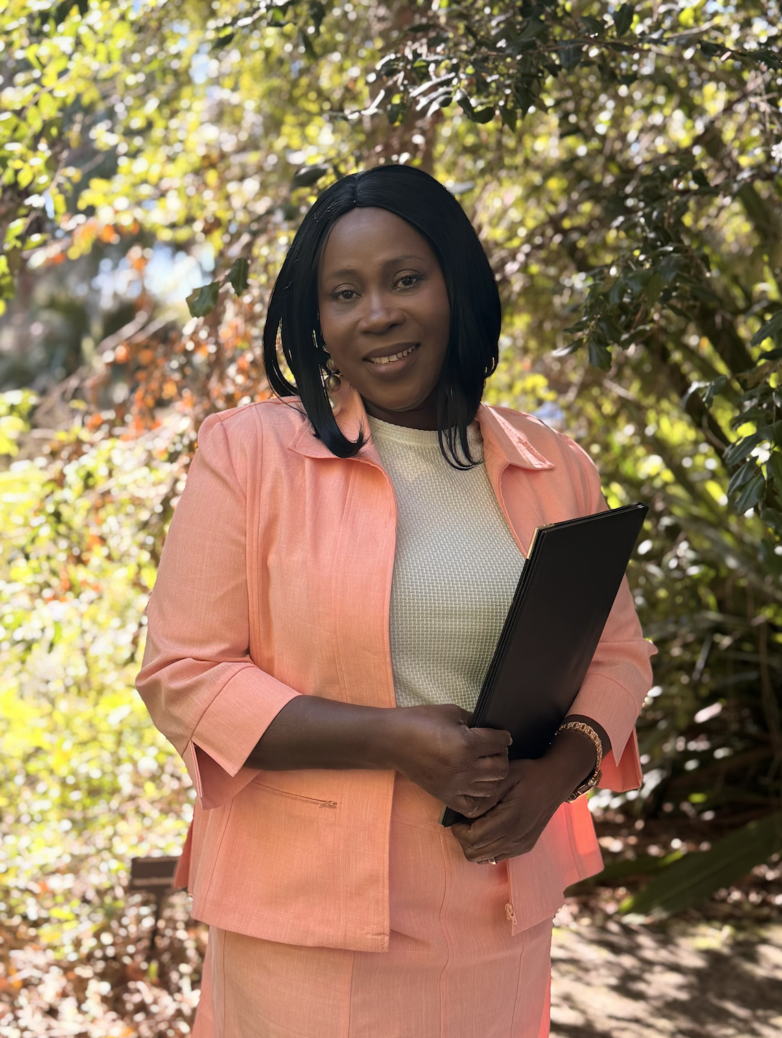 A woman in a peach-colored blazer and skirt holding a black folder, standing outdoors with green and brown leafy trees in the background.