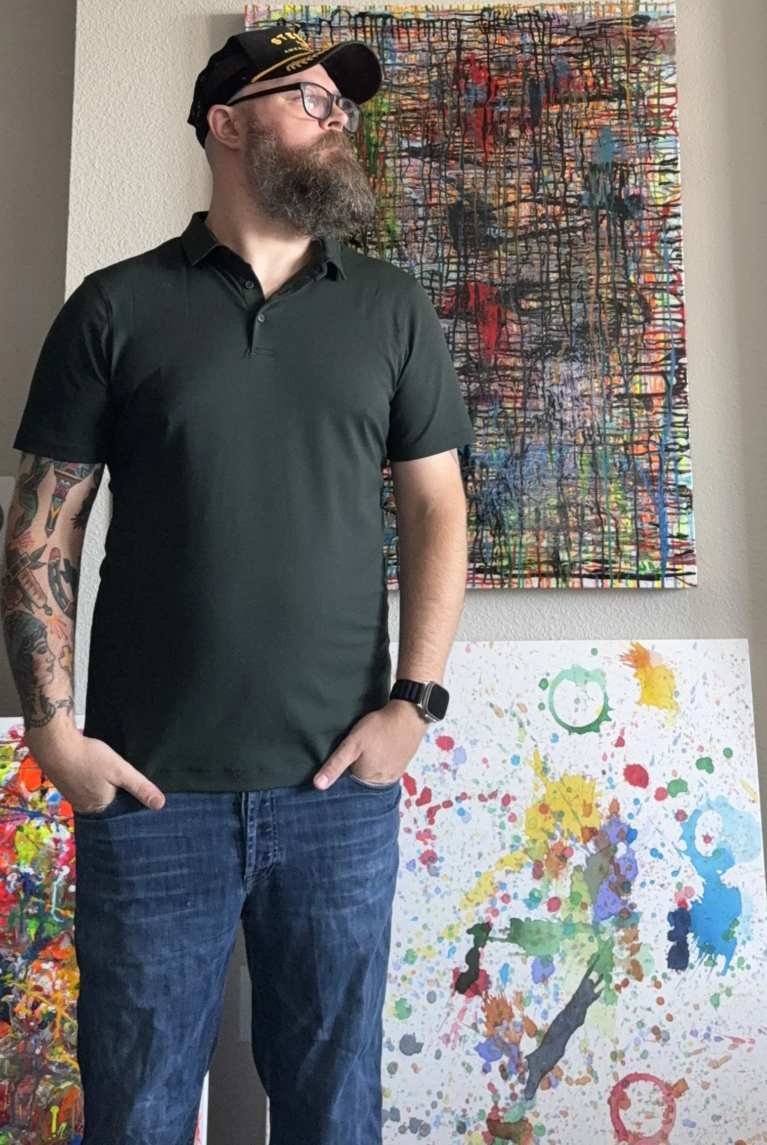 Josh Allen - Artist - with a beard, glasses, and a black cap stands in front of abstract splatter and drip paintings, wearing a black polo shirt and jeans.