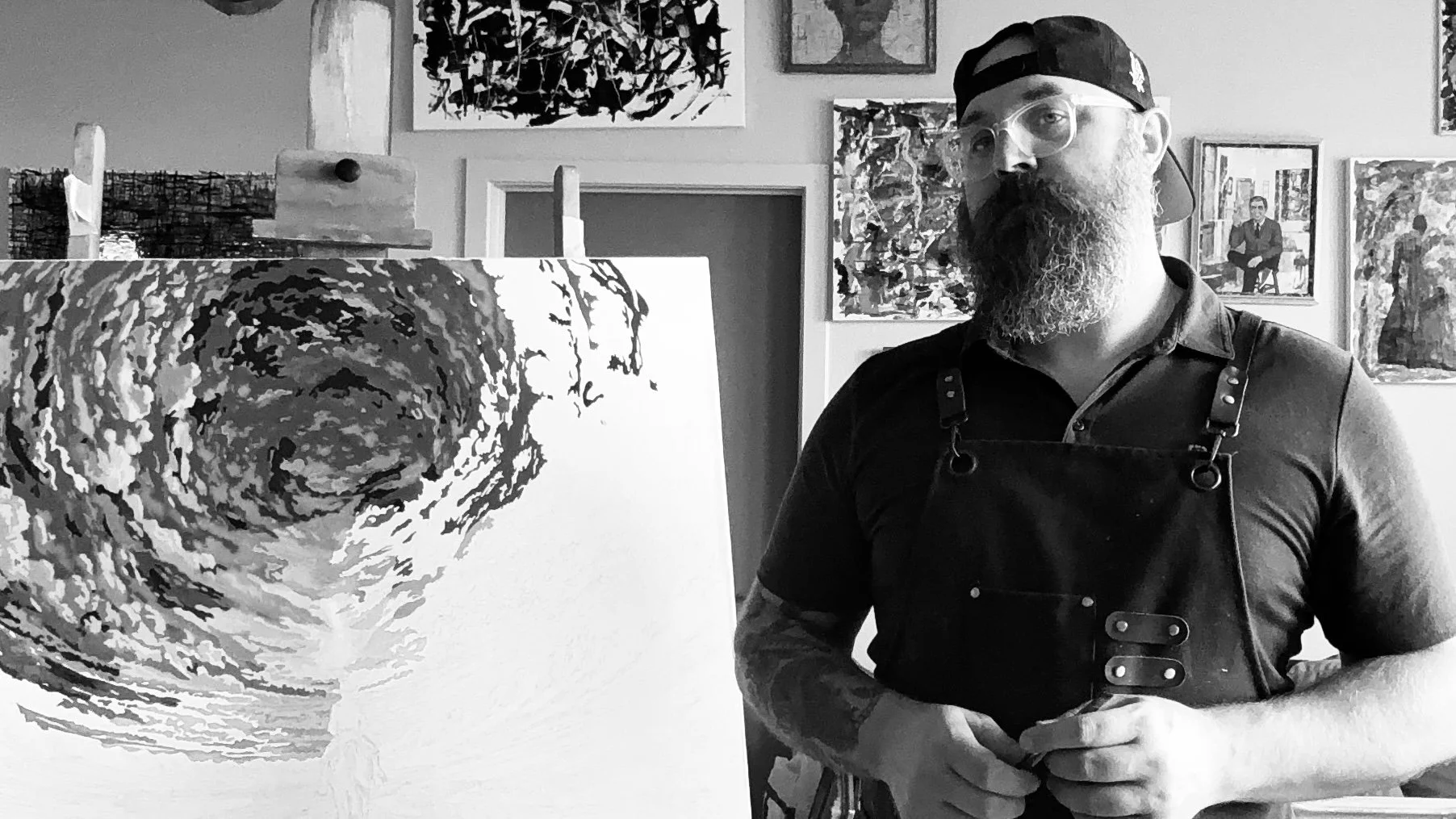 Josh Allen - Artist -with a beard, glasses, and a backward baseball cap stands in an art studio, wearing a leather apron. He is next to an abstract swirling black-and-white painting on an easel. The background has various framed artworks on the wall.