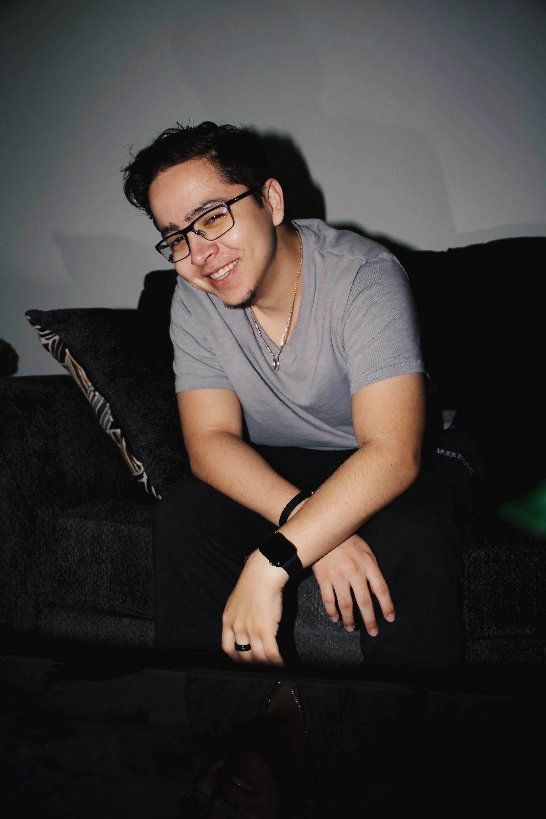 A young man with glasses smiling, wearing a gray t-shirt, sitting on a black couch with a pillow, in a dimly lit room.