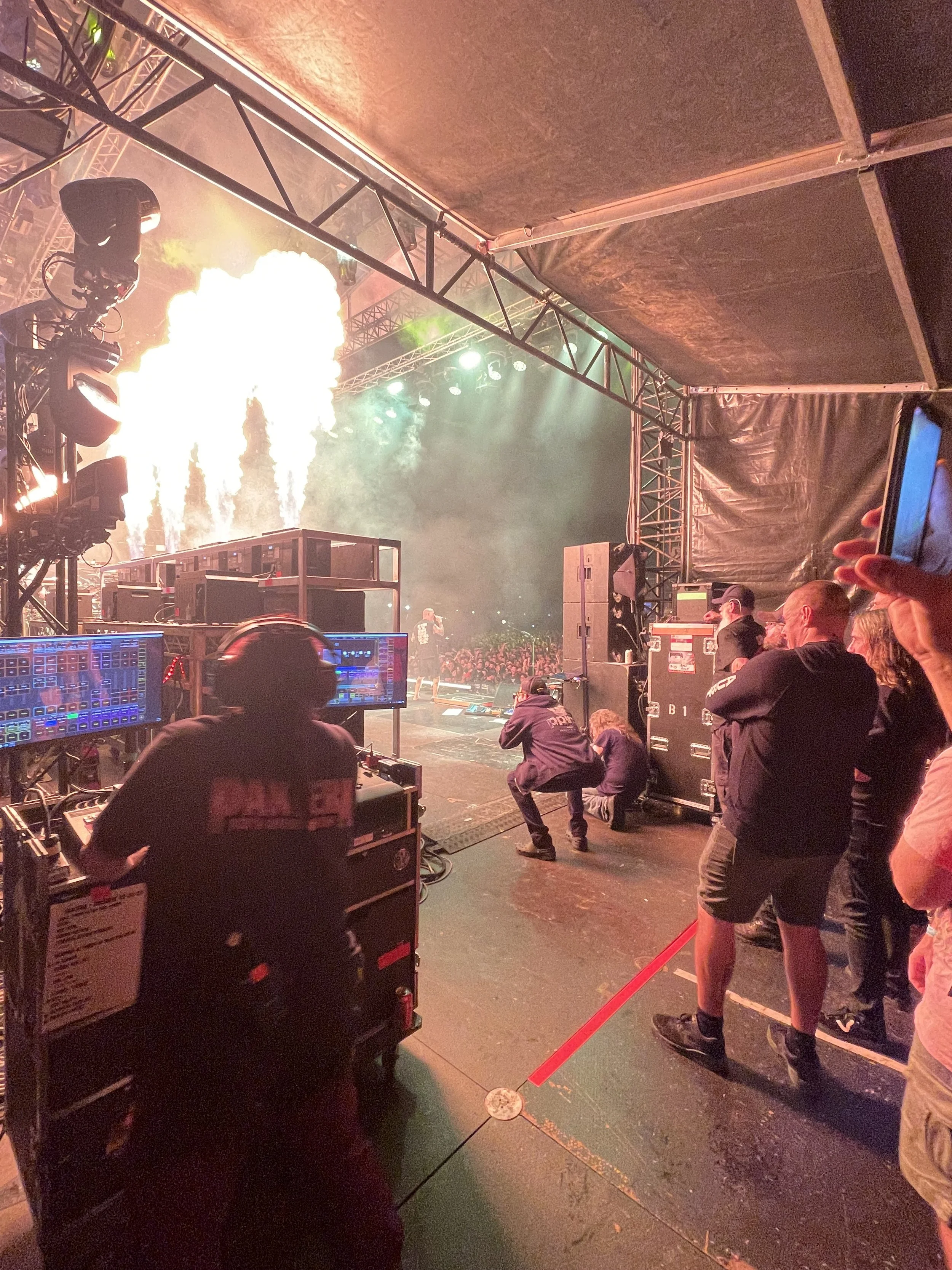 View from behind the stage at a concert, showing a crowd and performers, with pyrotechnics and colorful lights. Equipment and crew members are visible in the foreground.